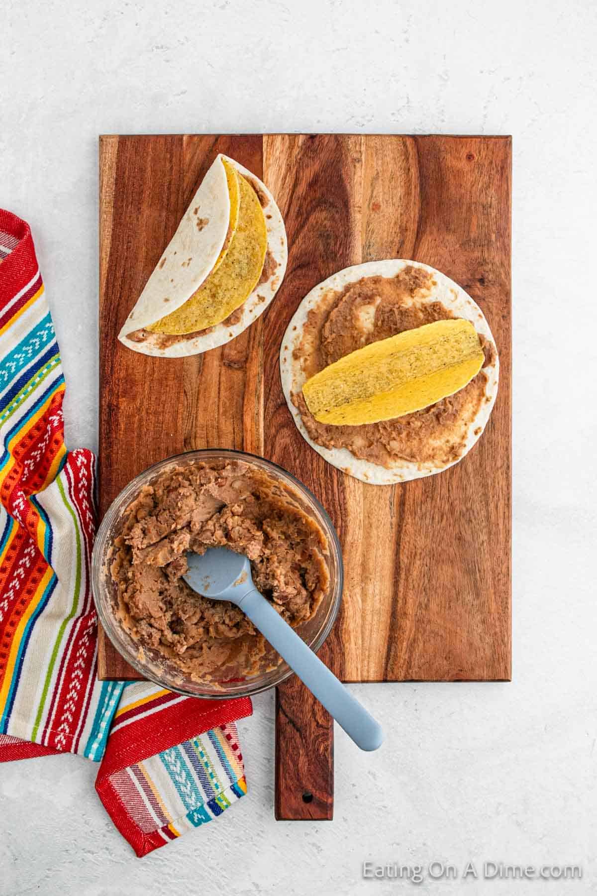 A wooden board showcases a Double Decker Taco Recipe with two tortillas spread with refried beans, topped with crispy taco shells. Nearby, a bowl of refried beans rests with a spoon, while a vibrant striped cloth adds flair to the light backdrop.