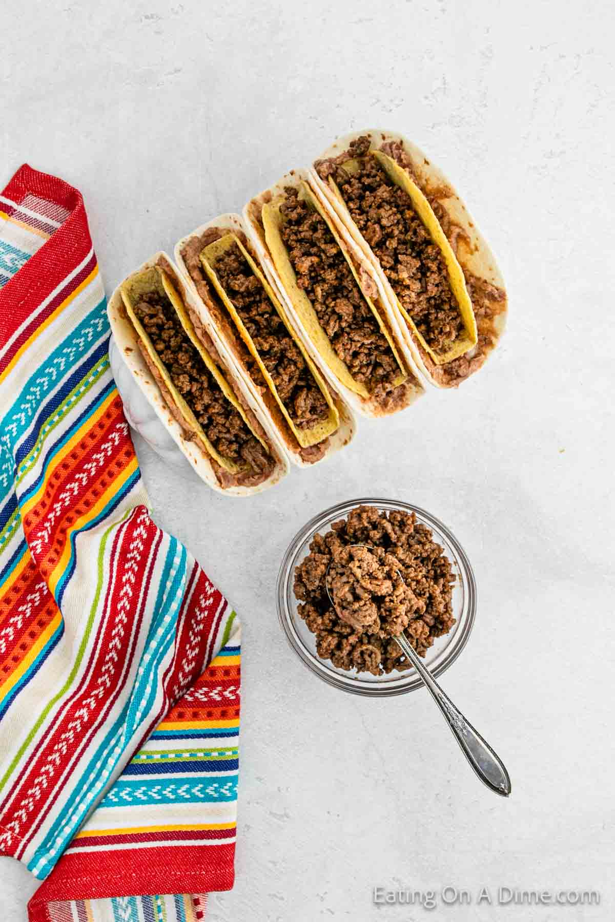 Five hard shell tacos filled with seasoned ground beef, reminiscent of a Double Decker Taco recipe, are arranged next to a colorful striped napkin. A small bowl of additional ground beef with a spoon is nearby.