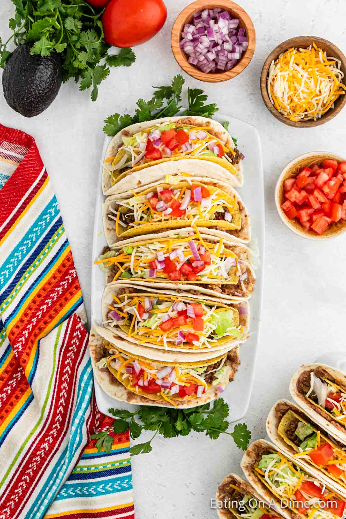 A platter of colorful double-decker tacos filled with various toppings like shredded cheese, diced tomatoes, and onions, surrounded by bowls of chopped vegetables and avocados on a white surface. A vibrant striped cloth sits beside this delightful taco recipe.