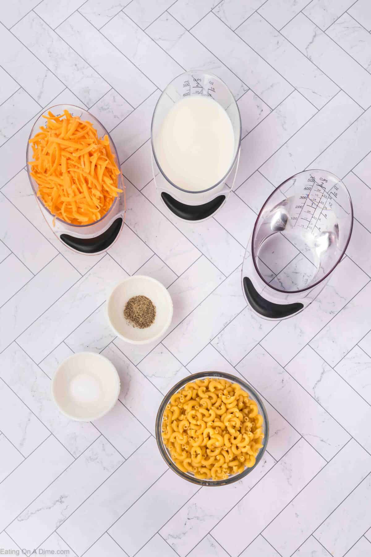 Ingredients for an easy homemade macaroni and cheese recipe laid out on a white tiled surface. Includes grated cheddar cheese, milk, pasta, salt, and pepper in separate bowls and measuring cups.