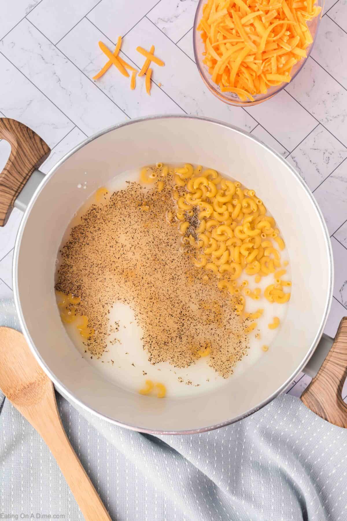 A pot on a white tiled countertop contains milk, elbow macaroni, and black pepper, ready to be cooked. Nearby, a bowl of shredded cheddar cheese and a wooden spoon await the next steps in this easy homemade macaroni and cheese recipe, with a gray cloth napkin completing the setup.