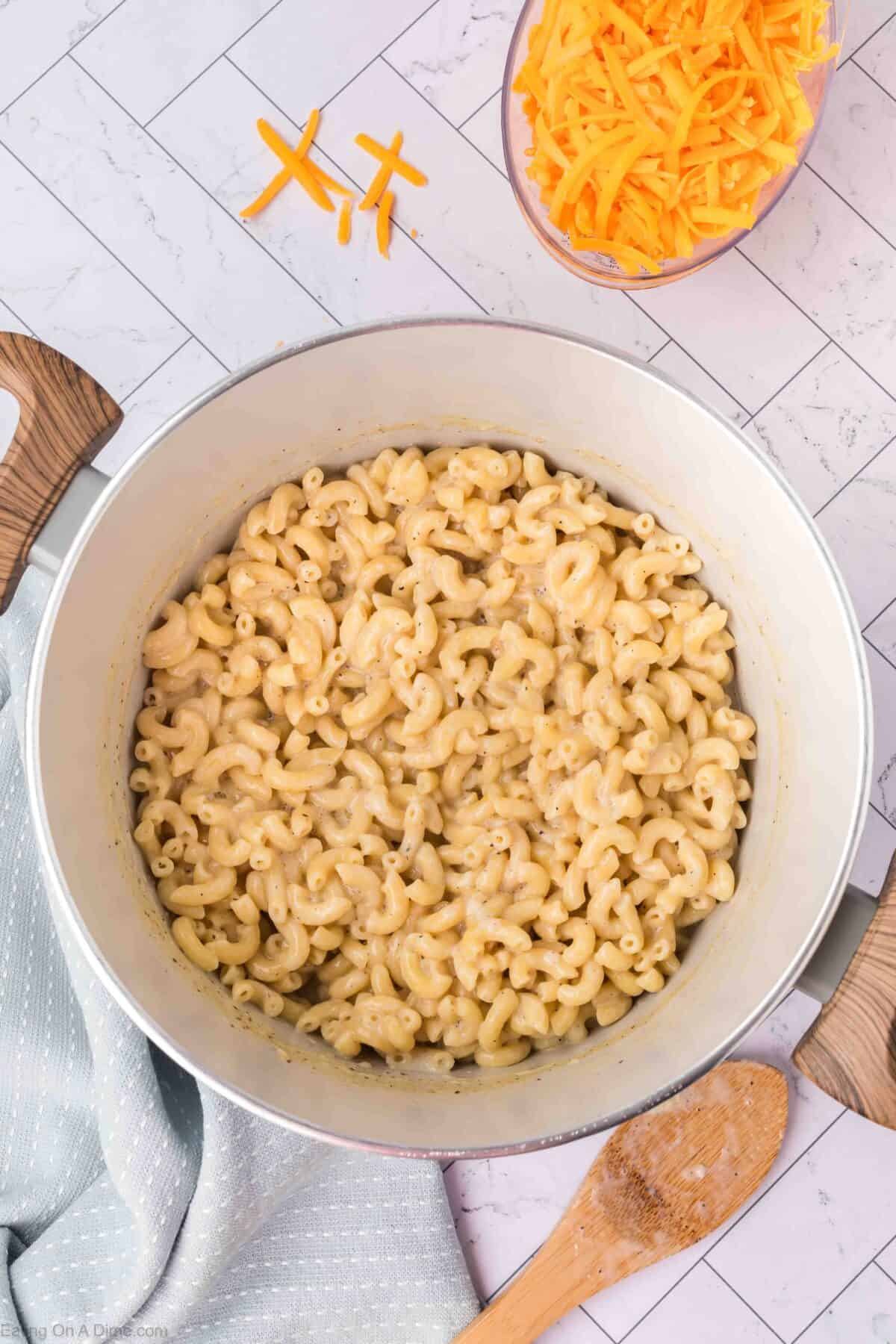A large pot of creamy macaroni and cheese, showcasing an easy homemade macaroni and cheese recipe, sits on a marble countertop. Next to it is a wooden spoon resting on a light blue cloth and a bowl filled with shredded cheddar cheese. A few pieces of cheddar are scattered on the counter.