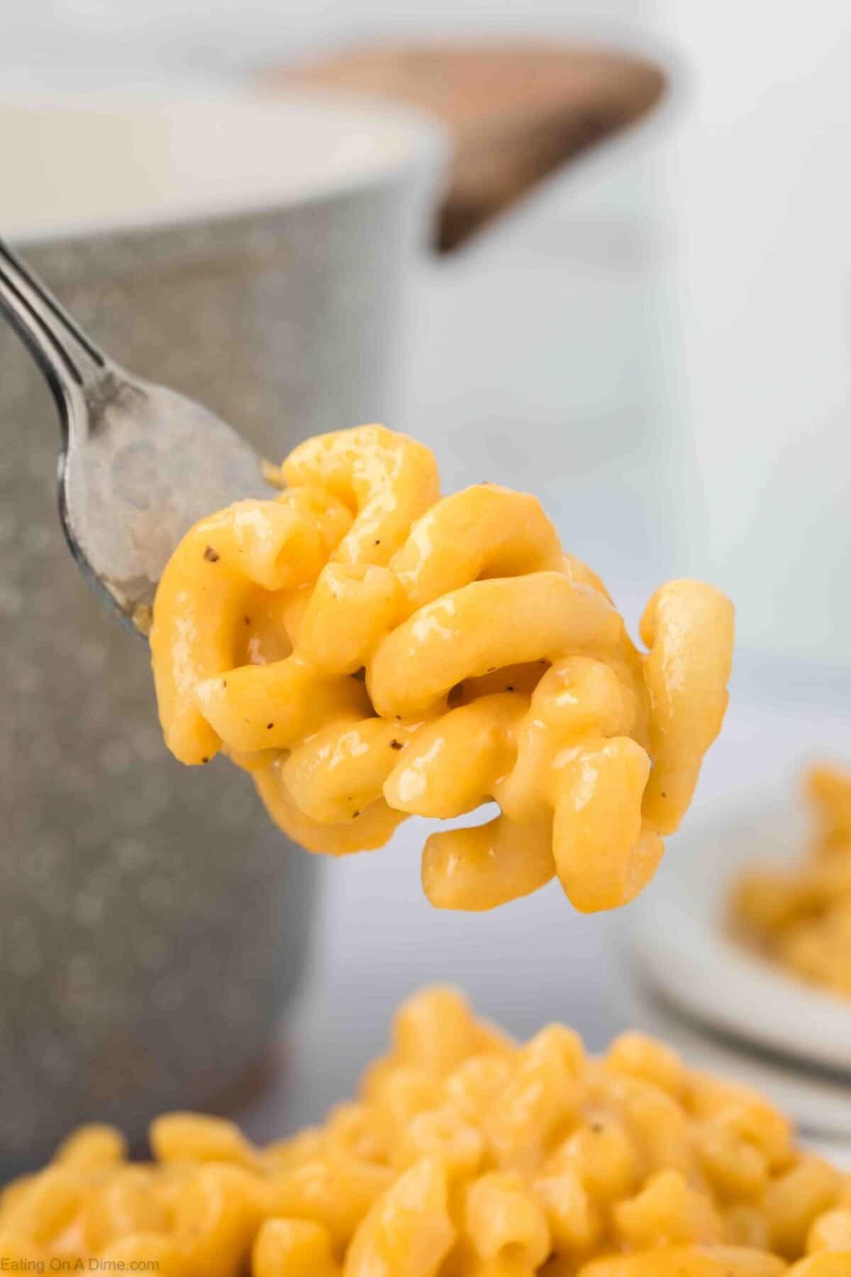 A close-up of a fork holding a bite of creamy, cheesy macaroni and cheese. The background shows a pot with more macaroni and cheese and a small plate with additional servings. The pasta is covered in a rich, yellow cheese sauce from an easy homemade macaroni and cheese recipe.