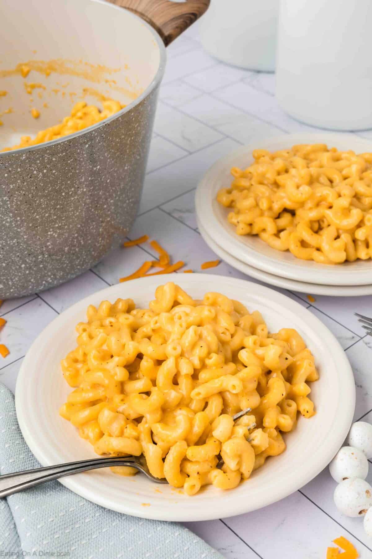 Two plates of creamy macaroni and cheese are set on a table next to a pot filled with more mac and cheese, all from an easy homemade macaroni and cheese recipe. The dishes are garnished with bits of shredded cheddar. Forks rest on the edge of each plate, ready for use.