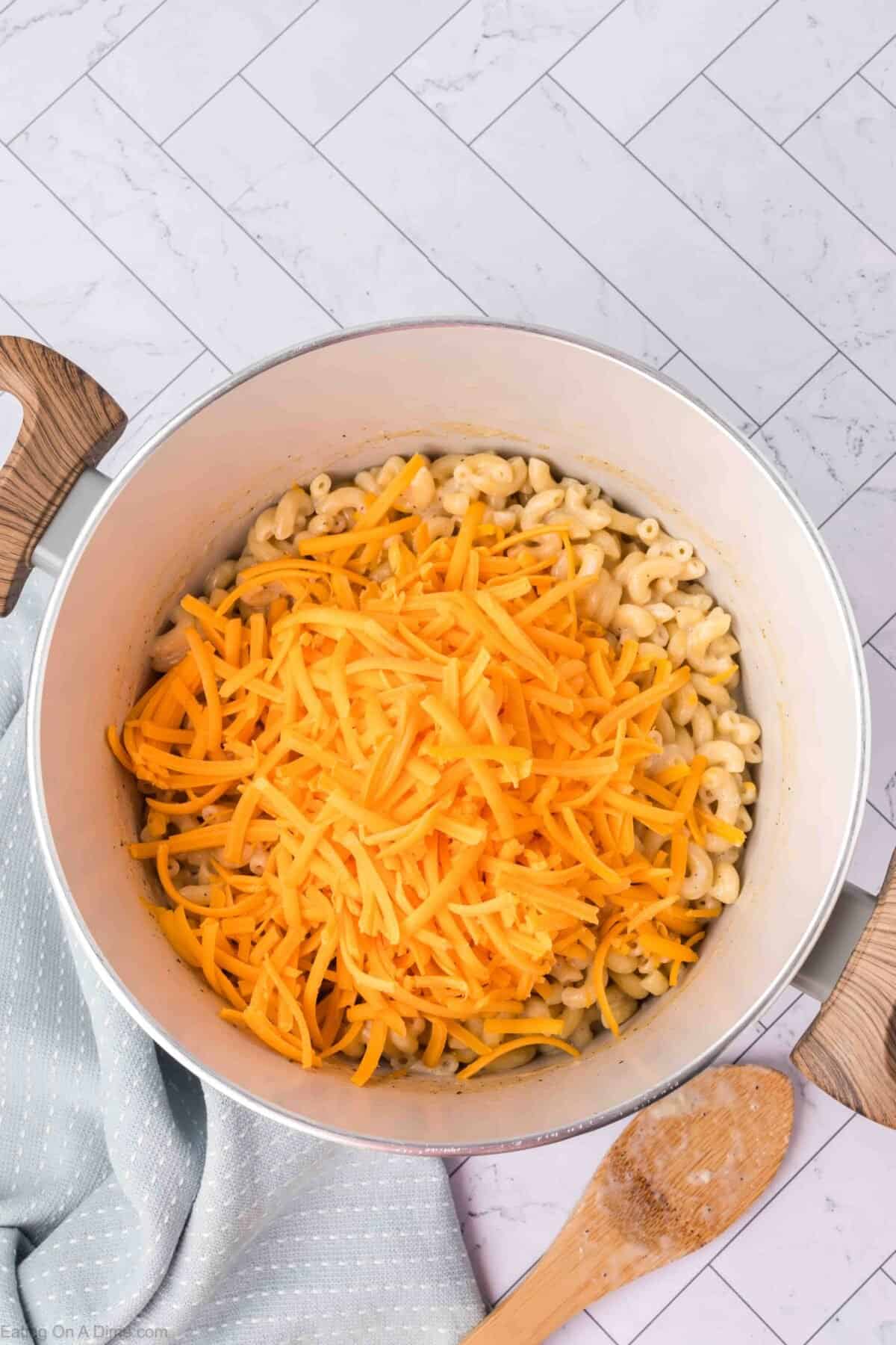 A large pot filled with cooked macaroni noodles topped with a generous amount of shredded cheddar cheese. The pot, part of an easy homemade macaroni and cheese recipe, is placed on a light blue kitchen towel, and a wooden spoon rests beside it on the tiled kitchen surface.