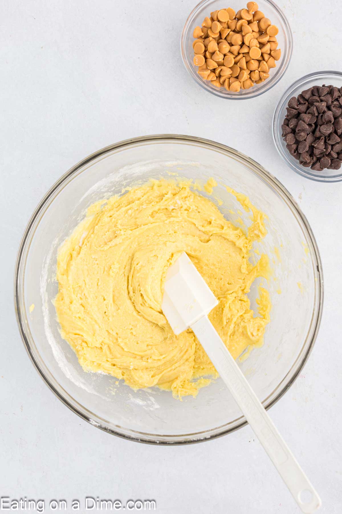 A glass bowl with cookie dough and a white spatula rests on a light surface, primed for making delicious Butterscotch Chocolate Chip Bars. Nearby, two small bowls await, one filled with butterscotch chips and the other brimming with chocolate chips.