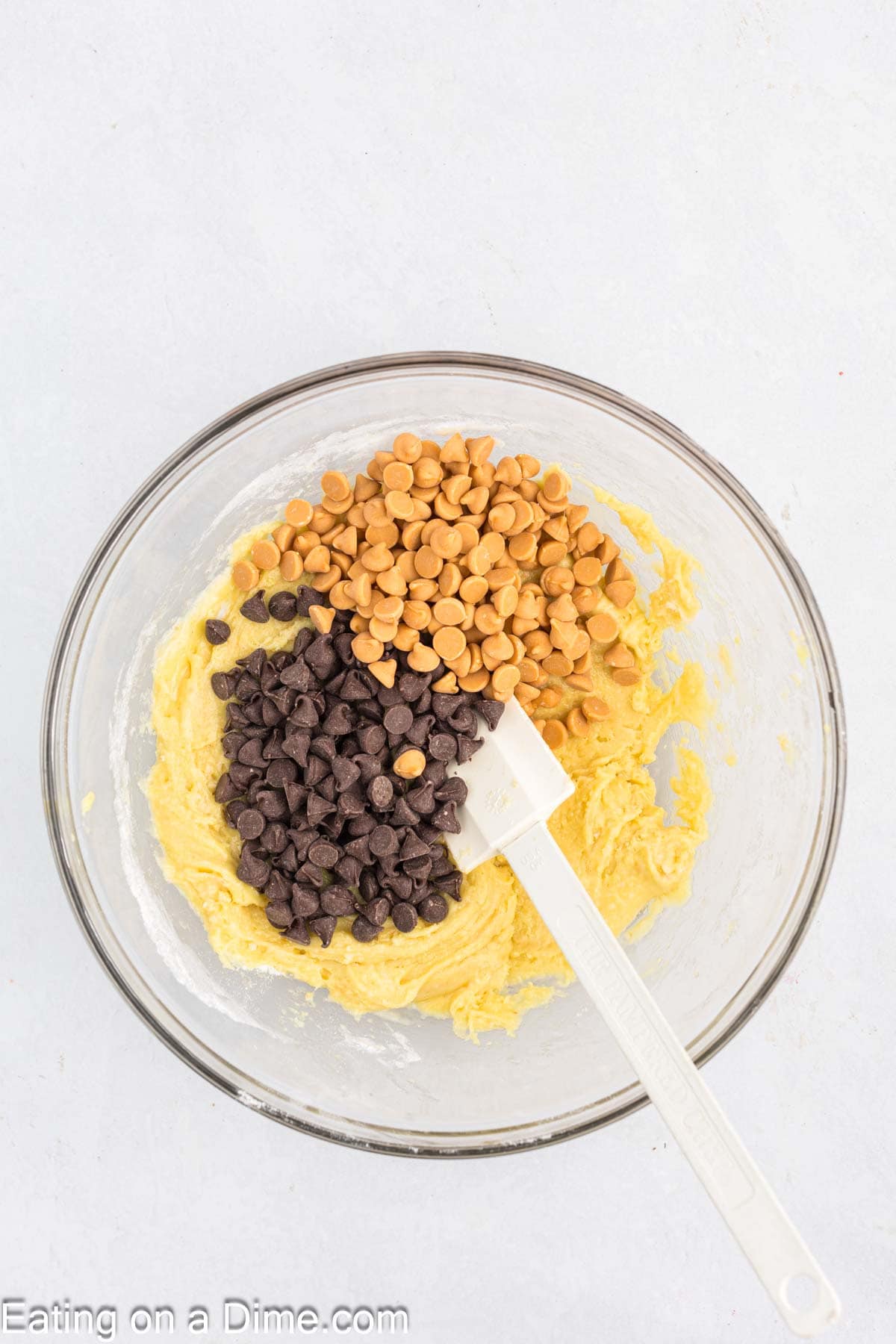 A glass bowl brimming with cookie dough, sprinkled with chocolate and butterscotch chips for delicious Butterscotch Chocolate Chip Bars. A white spatula rests in the mixture, all set against a light, textured backdrop.