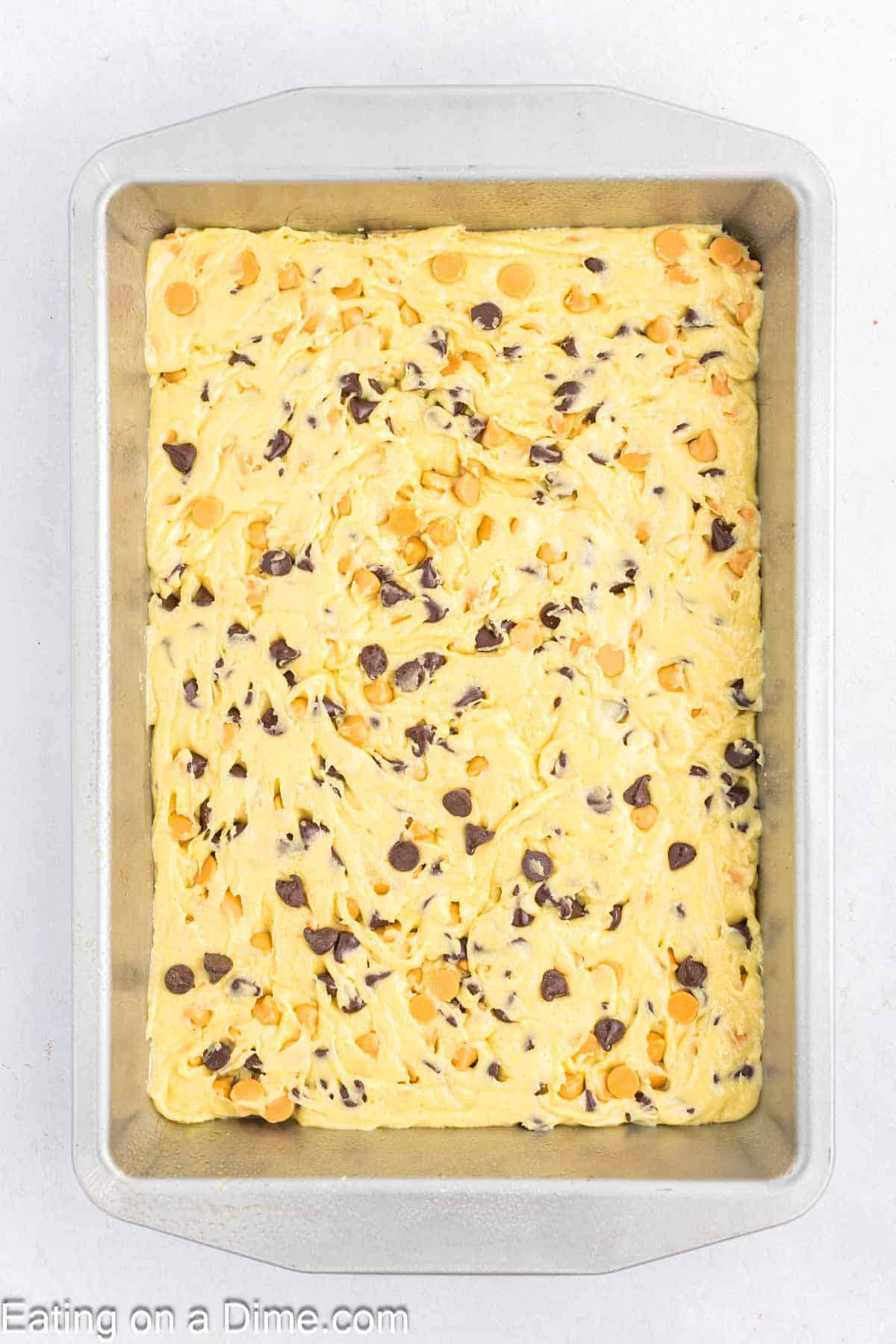 A rectangular baking pan brims with unbaked yellow cookie dough, perfectly mixed with chocolate and butterscotch chips. This delightful blend is evenly spread, primed to transform into delectable Butterscotch Chocolate Chip Bars.