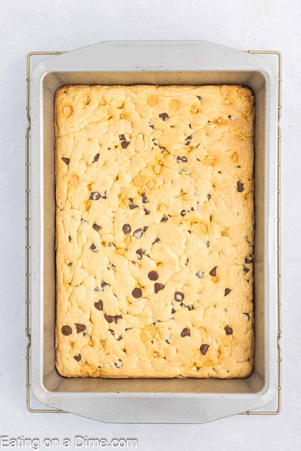 A baked tray of butterscotch chocolate chip bars sits in a rectangular metal pan, with a golden-brown top and scattered chocolate chips. The pan rests gracefully on a light gray surface.