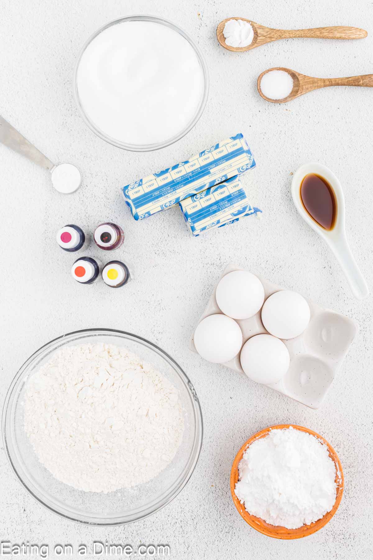 An assortment of baking ingredients is arranged on a white surface, perfect for creating Fall Leaf Sugar Cookies. These include a bowl of flour, eggs, butter sticks, a bowl of sugar, powdered sugar, a small bowl of vanilla extract, colored food dyes, and small wooden spoons with baking powder and salt. Text at the bottom reads "Eating on a Dime.com.