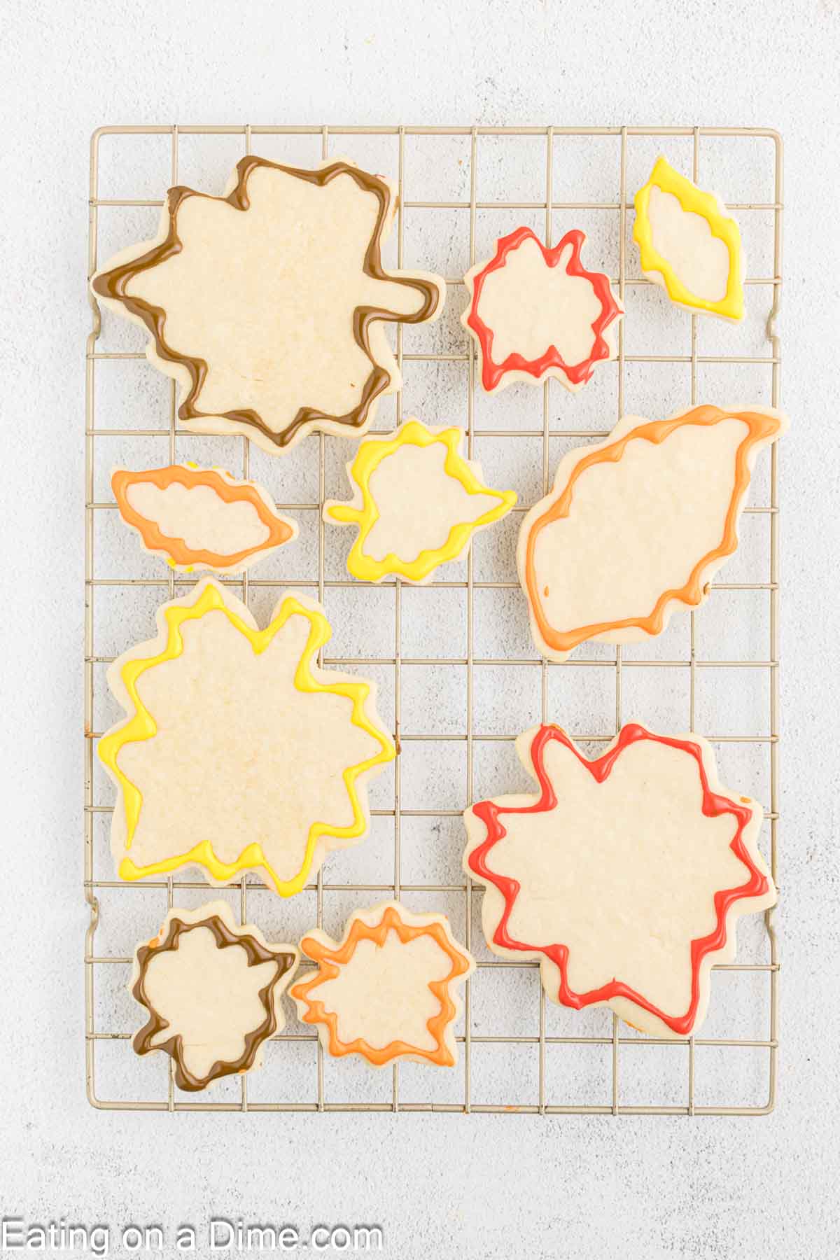 Assorted Fall Leaf Sugar Cookies with colorful borders in orange, yellow, red, and brown are laid out on a cooling rack. The cookies are in various sizes and shapes, resembling autumn leaves. The background is a light textured surface.