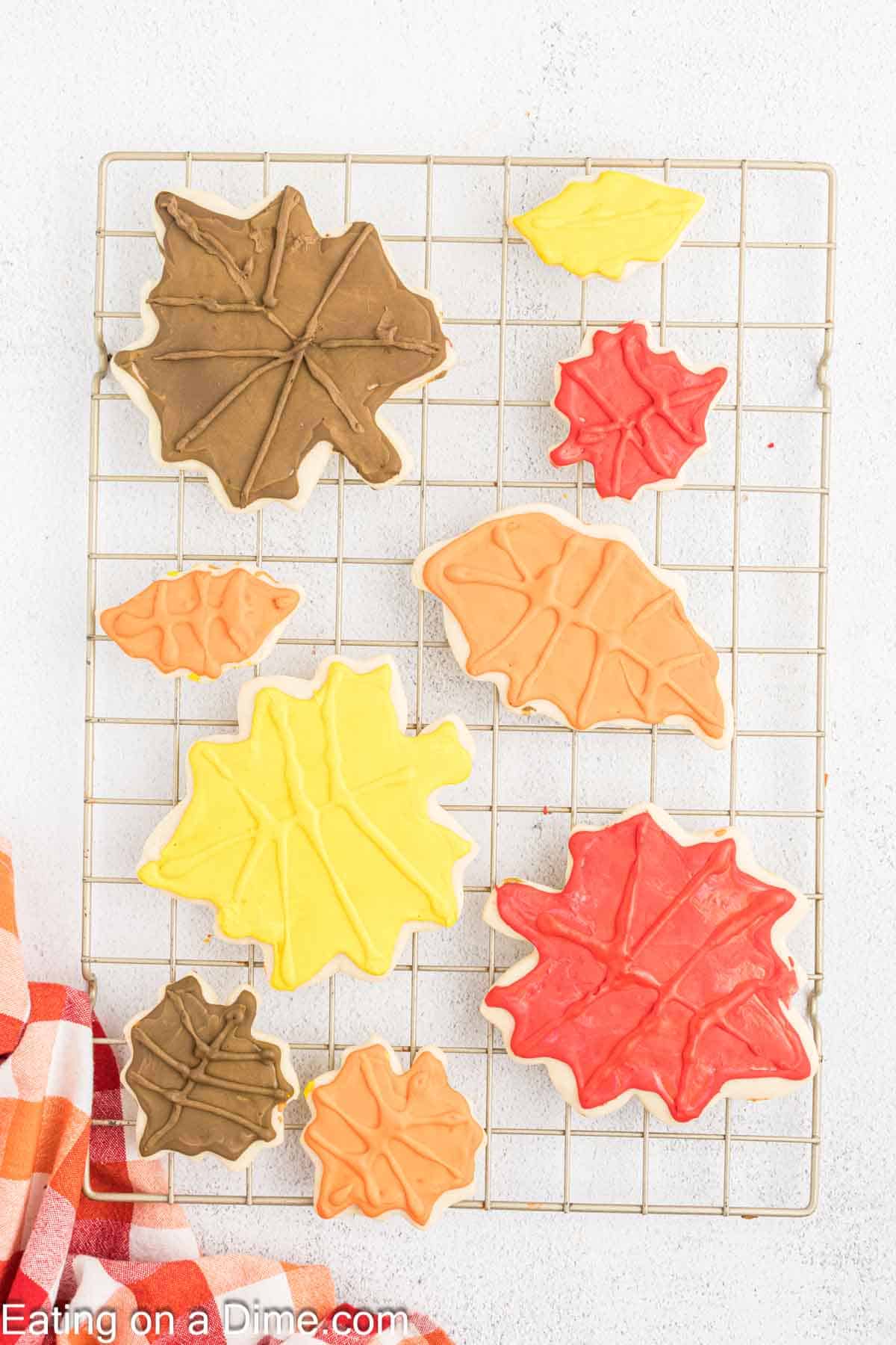 A cooling rack holds seven fall leaf sugar cookies decorated with autumn-colored icing—brown, yellow, orange, and red. A red and orange cloth is partially visible in the bottom left corner. The background is a white textured surface.