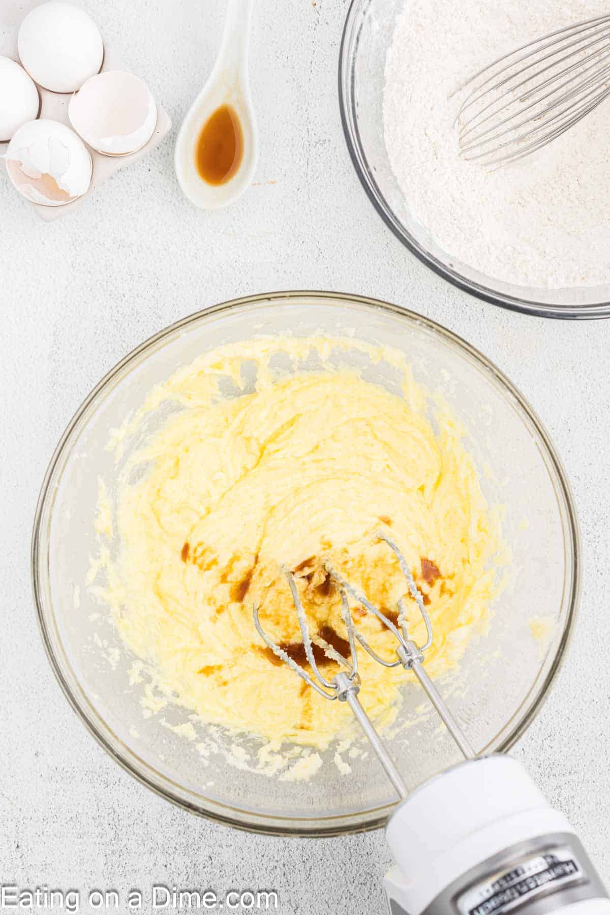 A mixing bowl filled with creamed butter and sugar, with a splash of vanilla being mixed in by a hand mixer. Surrounding the bowl are broken egg shells, a spoon with vanilla, and another bowl containing flour and a whisk. Text says, "Eating on a Dime.com." Perfect for making Fall Leaf Sugar Cookies!