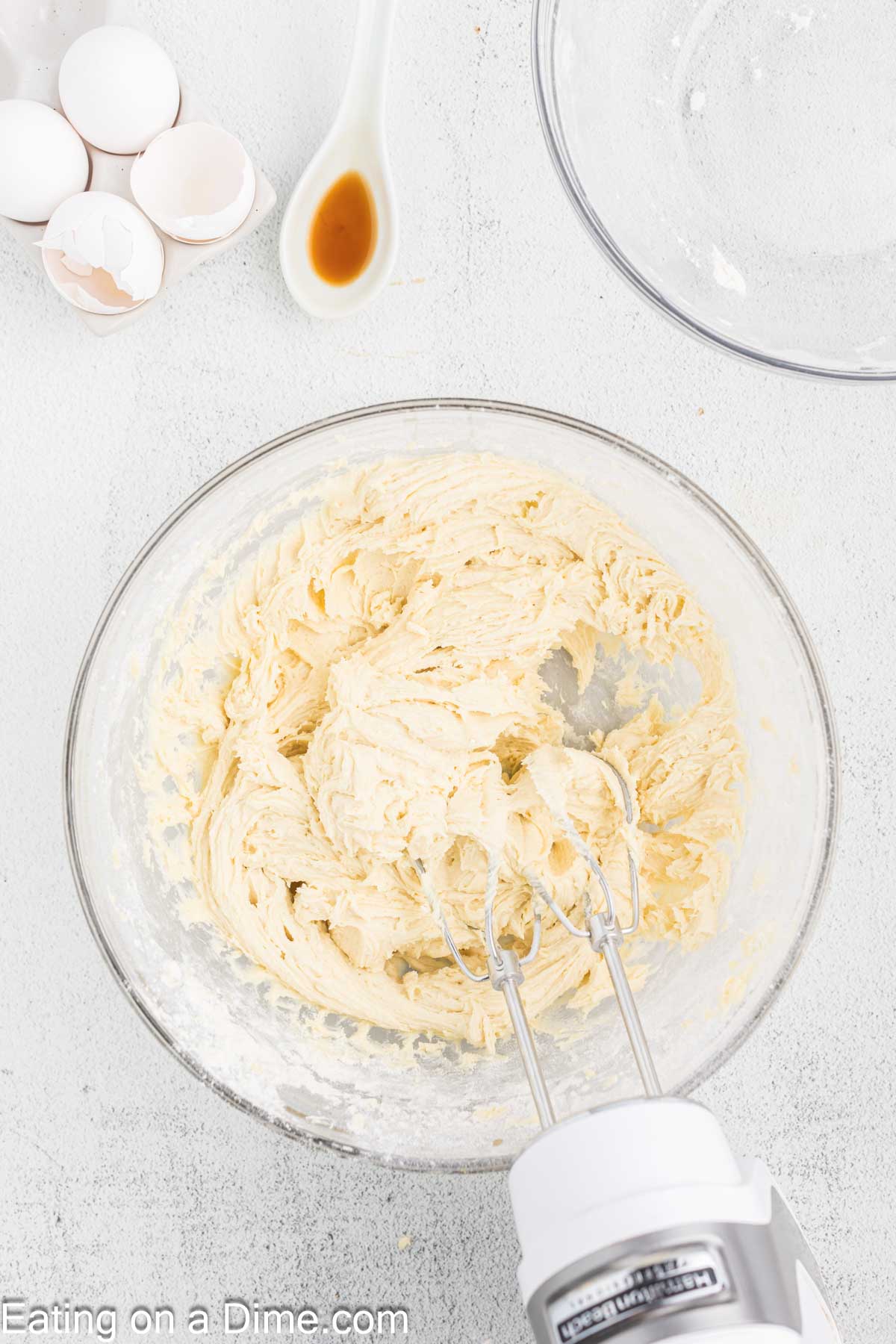 A mixing bowl containing a creamy batter is being blended with a hand mixer. To the side, there are cracked eggshells, a spoon with vanilla extract, and an empty clear bowl—preparations for delightful Fall Leaf Sugar Cookies. The clean white surface adds a striking contrast. Text reads: "Eating on a Dime.com.