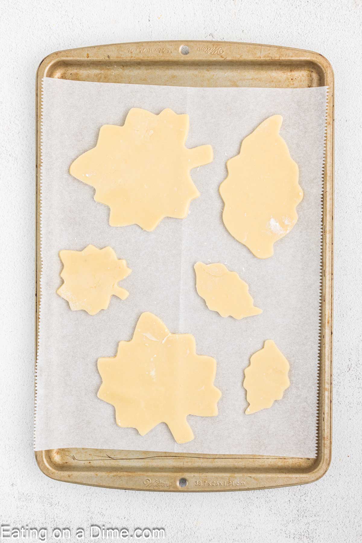 A baking sheet lined with parchment paper holds six unbaked pie crust shapes resembling various sizes of autumn leaves, ready for baking. These fall leaf sugar cookies capture the essence of the season perfectly.