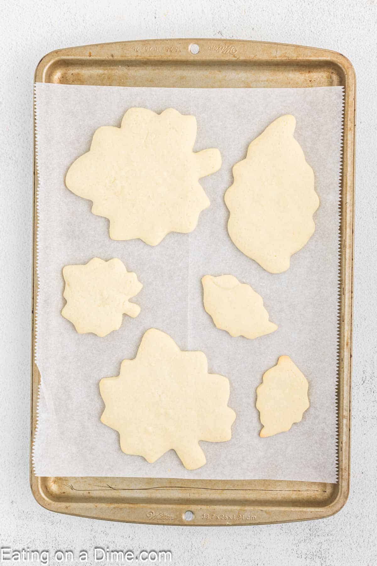 A baking tray lined with parchment paper holds six raw, leaf-shaped cookie dough cutouts. These Fall Leaf Sugar Cookies vary in size and type of leaves. The parchment paper edges are visible, and the background is a light, textured surface.