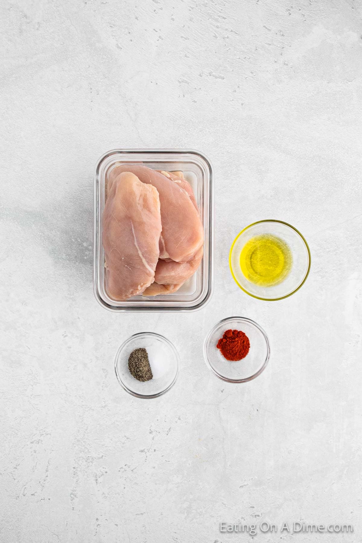 Chicken breast in a glass container with bowls of oil, paprika, salt and pepper