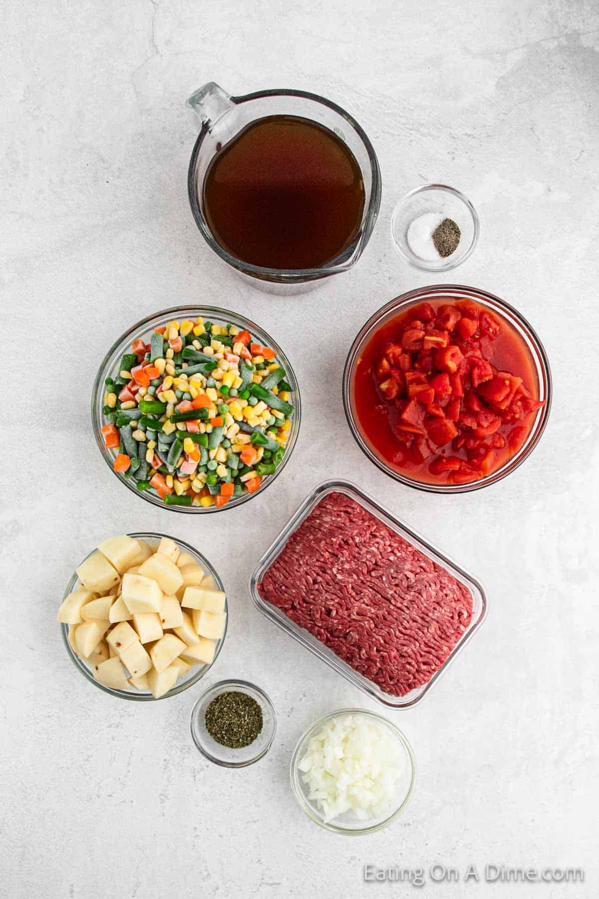 Top view of ingredients arranged on a white surface for making an Instant Pot Beef Vegetable Soup Recipe. The setup includes a large glass measuring cup with brown liquid, a small bowl of black pepper, canned diced tomatoes, mixed frozen vegetables, diced potatoes, ground beef, chopped onions, and dried herbs.