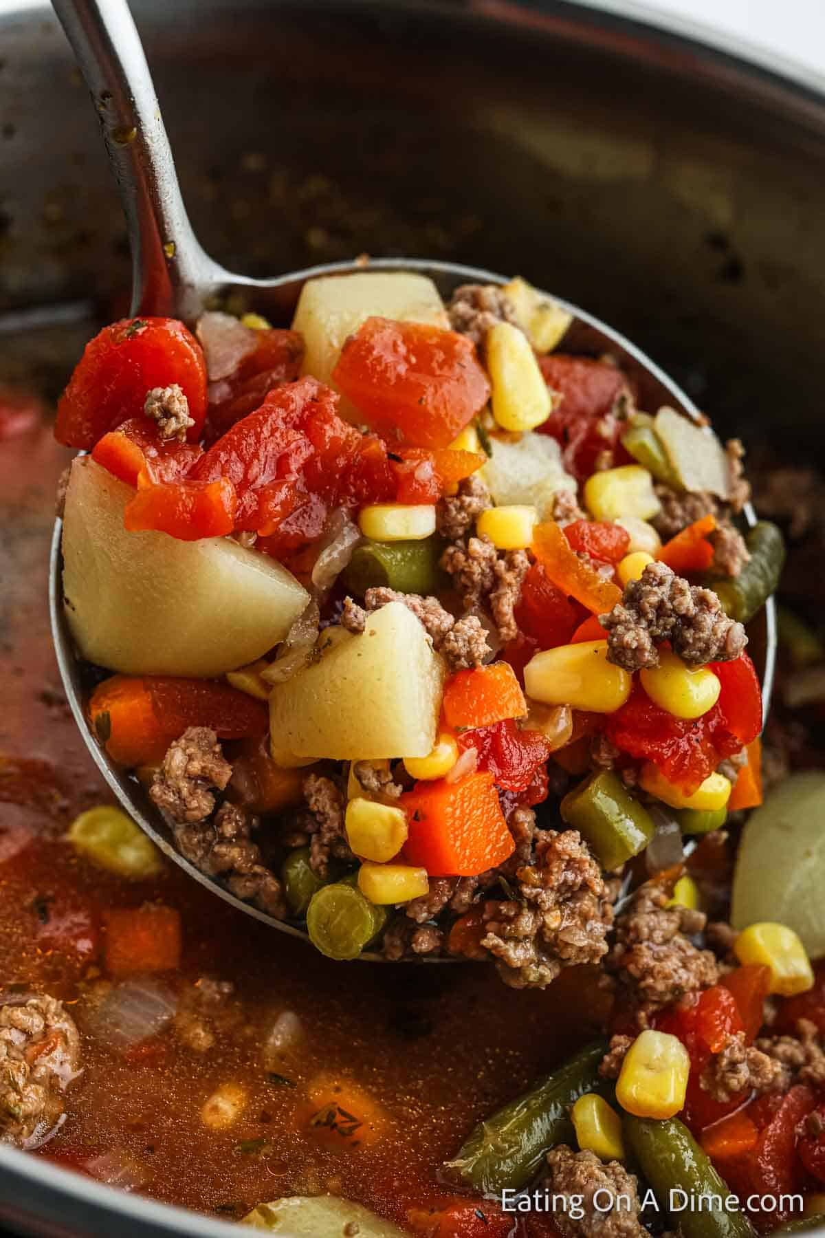 A ladle filled with hearty Instant Pot beef vegetable soup containing chunks of potato, ground meat, diced tomatoes, corn, carrots, green beans, and peas hovers above a pot of the soup. The background shows the soup pot with the same vibrant mixture of ingredients.