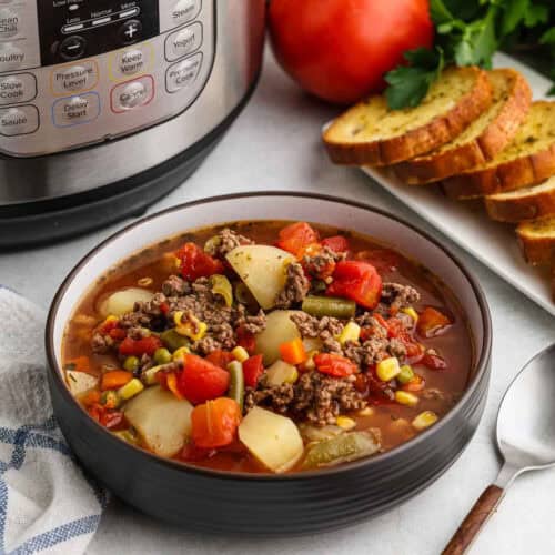 A bowl of hearty Instant Pot Beef Vegetable Soup sits on a table next to a plate with slices of garlic bread and a fresh tomato. The soup is filled with ground beef, potatoes, corn, green beans, and diced tomatoes. A spoon rests nearby for an inviting meal.