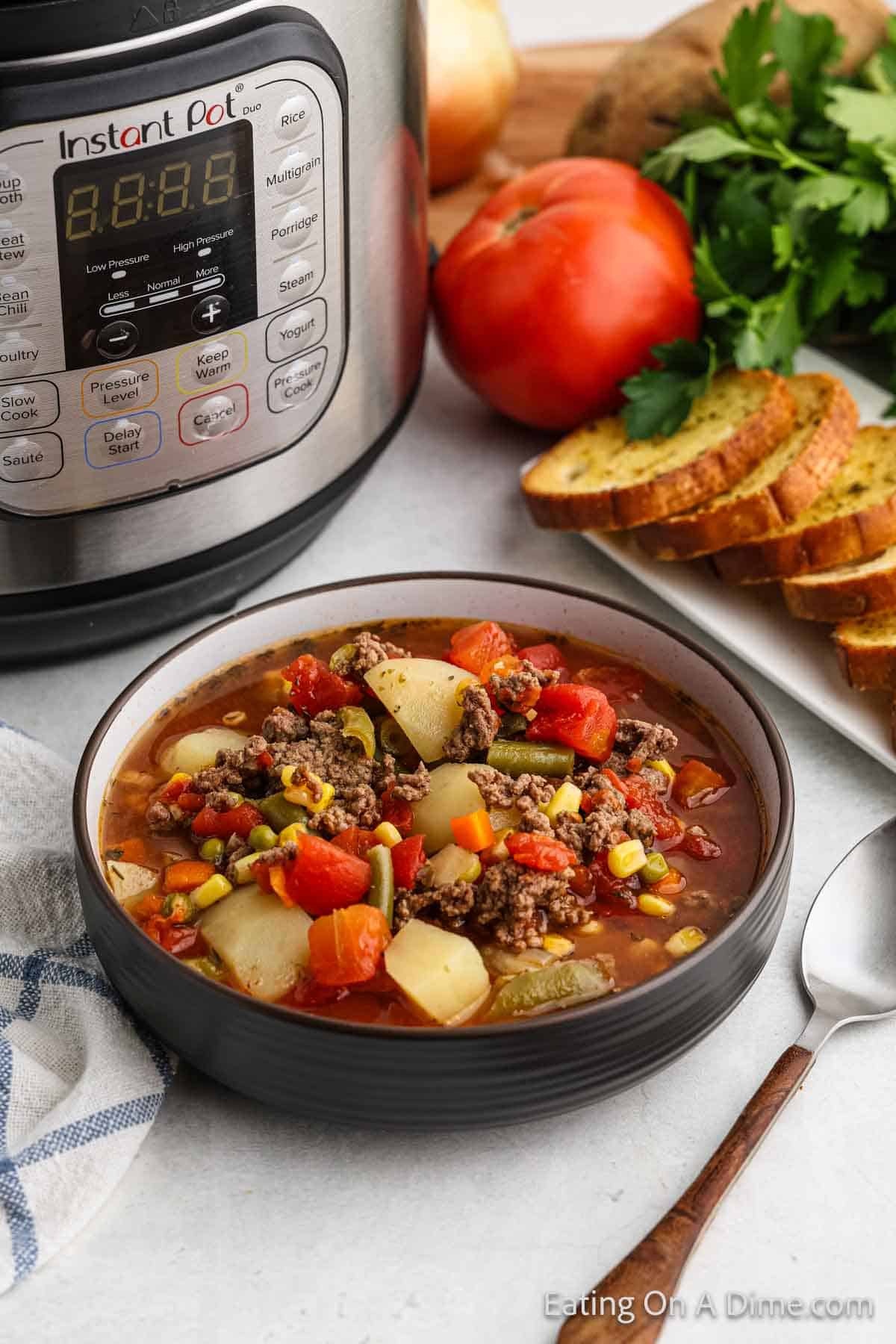 A bowl of hearty Instant Pot Beef Vegetable Soup with diced vegetables and ground beef is placed in front of an Instant Pot. Fresh parsley, sliced bread, a red tomato, and a spoon are on the side, suggesting a cozy homemade meal perfect for following an easy recipe.