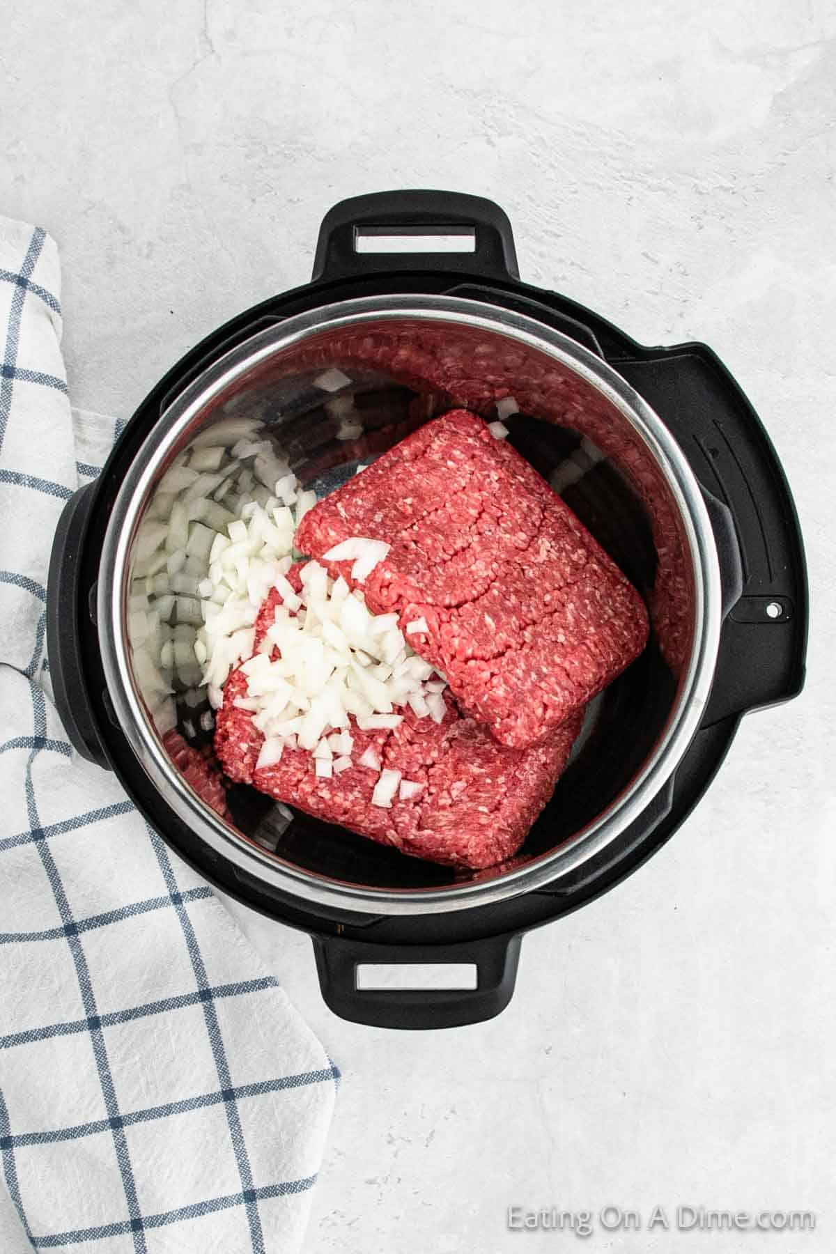 An overhead view of a pressure cooker pot containing two blocks of ground beef and chopped onions, perfect for starting an Instant Pot Beef Vegetable Soup Recipe. The pot rests on a light-colored countertop next to a white and blue checkered cloth, with a watermark that reads "Eating On A Dime.com".