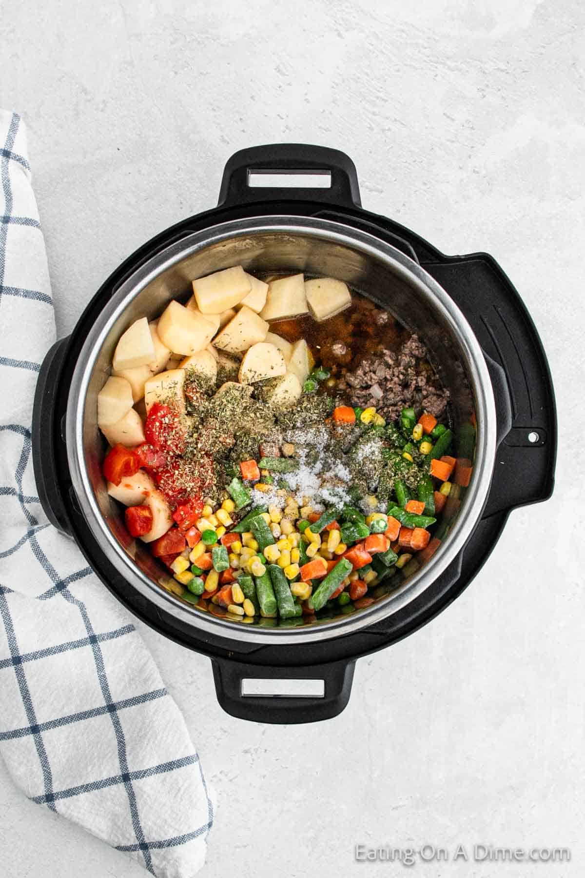 An opened Instant Pot filled with ingredients for a beef vegetable soup recipe. It contains diced potatoes, tomatoes, ground beef, mixed vegetables including carrots, green beans, corn, and peas, all topped with herbs and seasoning. A checkered cloth is nearby.