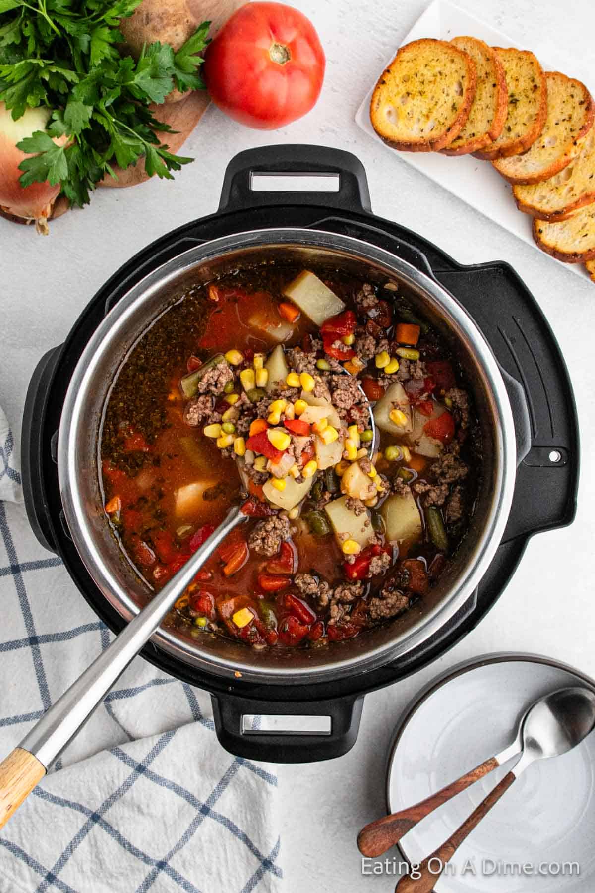 A top view of a pot filled with instant pot beef vegetable soup. The hearty soup contains ground beef, potatoes, corn, carrots, and beans in a savory broth. Surrounding the pot are slices of toasted bread, fresh parsley, a tomato, and a striped kitchen towel.