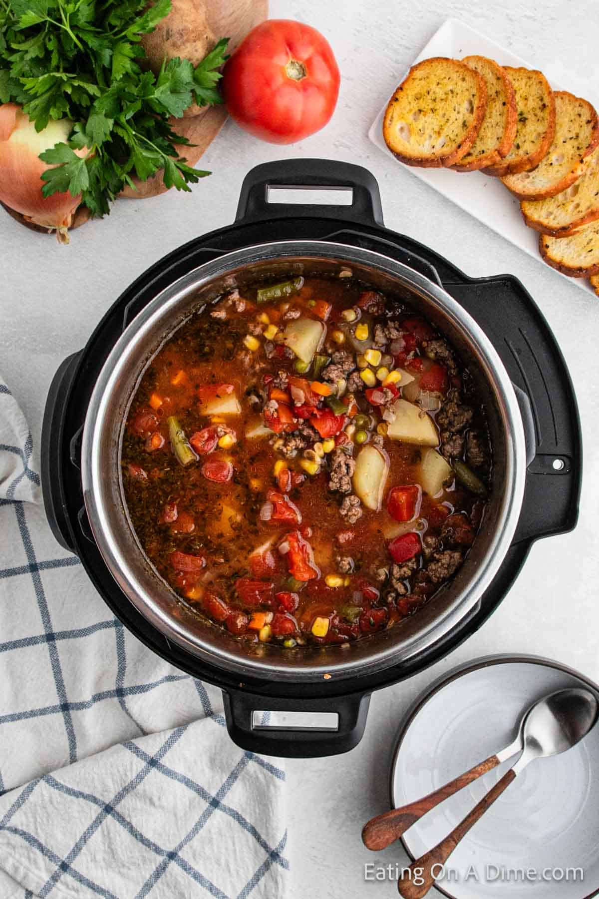 An overhead view of a hearty Instant Pot Beef Vegetable Soup in a black and steel slow cooker. The soup contains vegetables like corn, potatoes, and tomatoes. Surrounding the slow cooker are slices of toasted bread, a whole tomato, parsley, and a striped kitchen towel with a bowl and spoon.