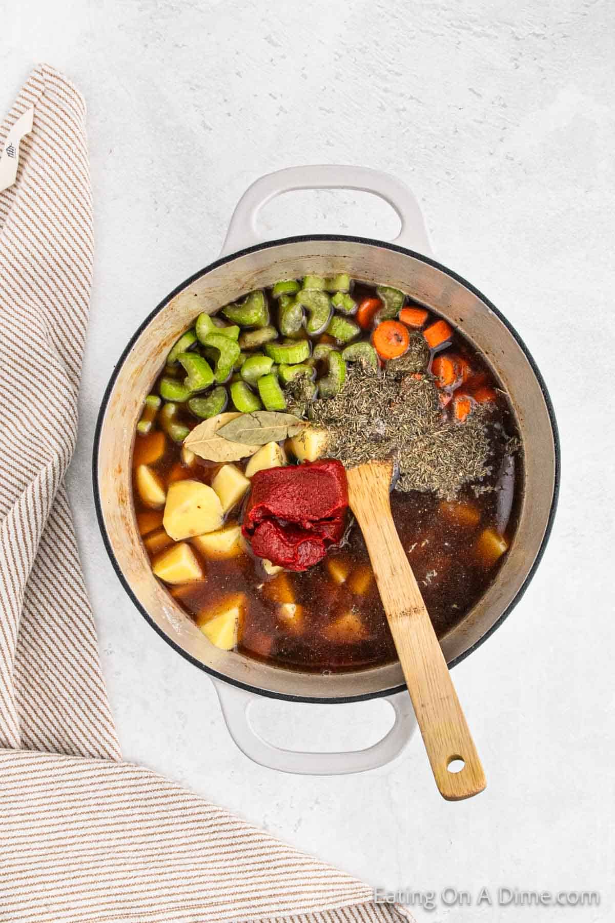 Adding broth, celery, carrots, potatoes, seasoning and tomato paste to large pot with wooden spoon