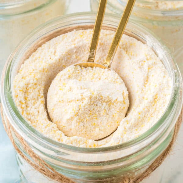 A close-up of a jar filled with cornmeal for your favorite cornbread mix recipe, with a golden spoon scooping some out. The jar is decorated with twine around its neck, and the cornmeal appears coarse and textured.