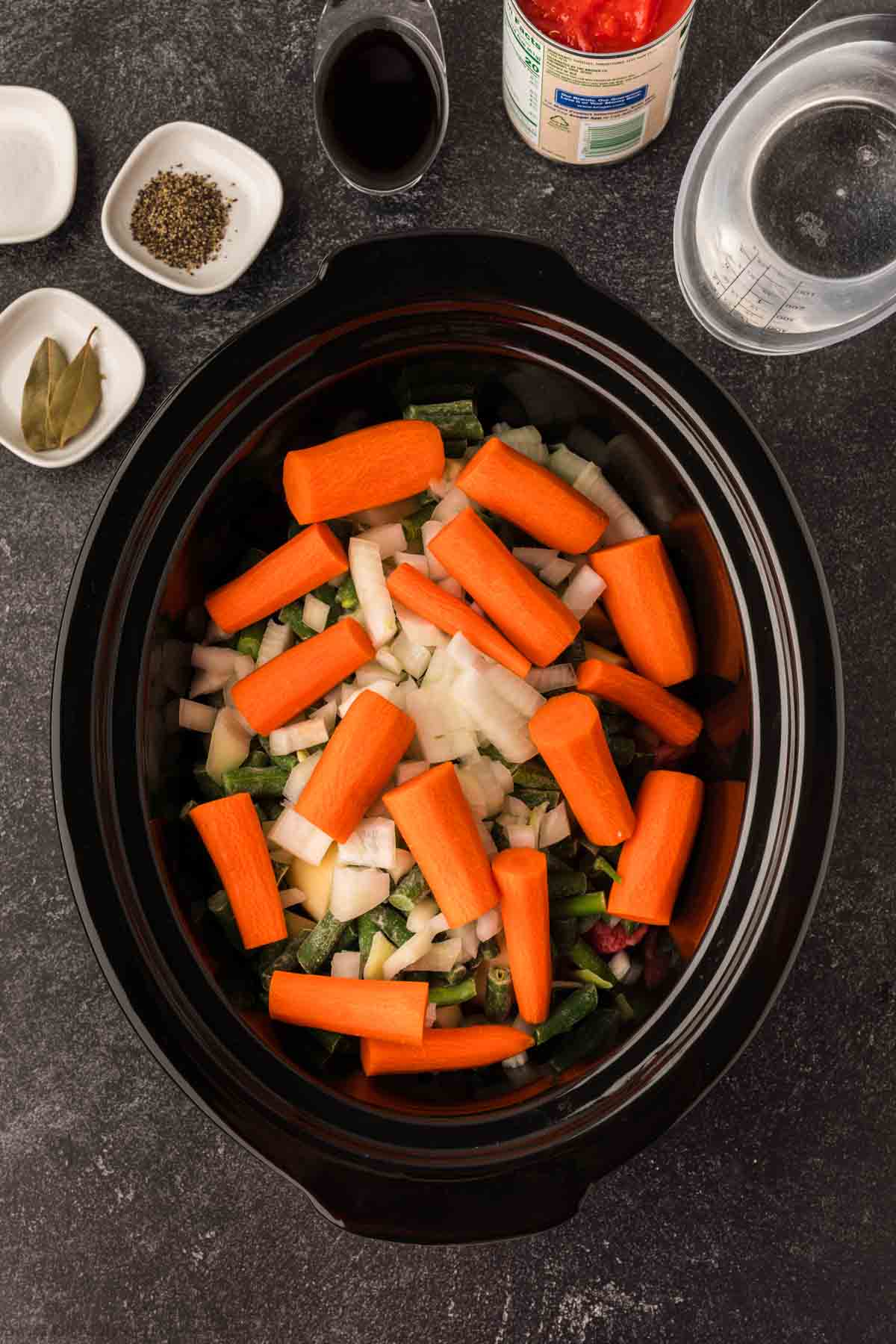 A slow cooker, perfect for a delicious Crock Pot Beef Stew recipe, is filled with chopped carrots, onions, and green peppers. Nearby sit small dishes of pepper and bay leaves, a can of tomatoes, a glass of water, and a bowl of dark sauce on the dark surface.