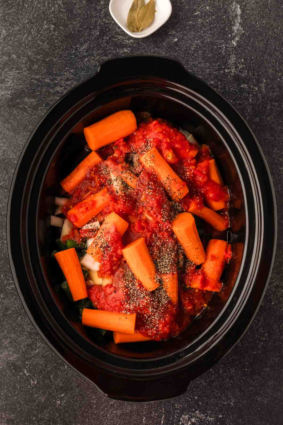 A slow cooker filled with fresh ingredients, including raw carrot sticks, diced tomatoes, spices, and additional vegetables for a hearty Crock Pot beef stew recipe. A small white dish with a bay leaf is placed nearby on the dark countertop.