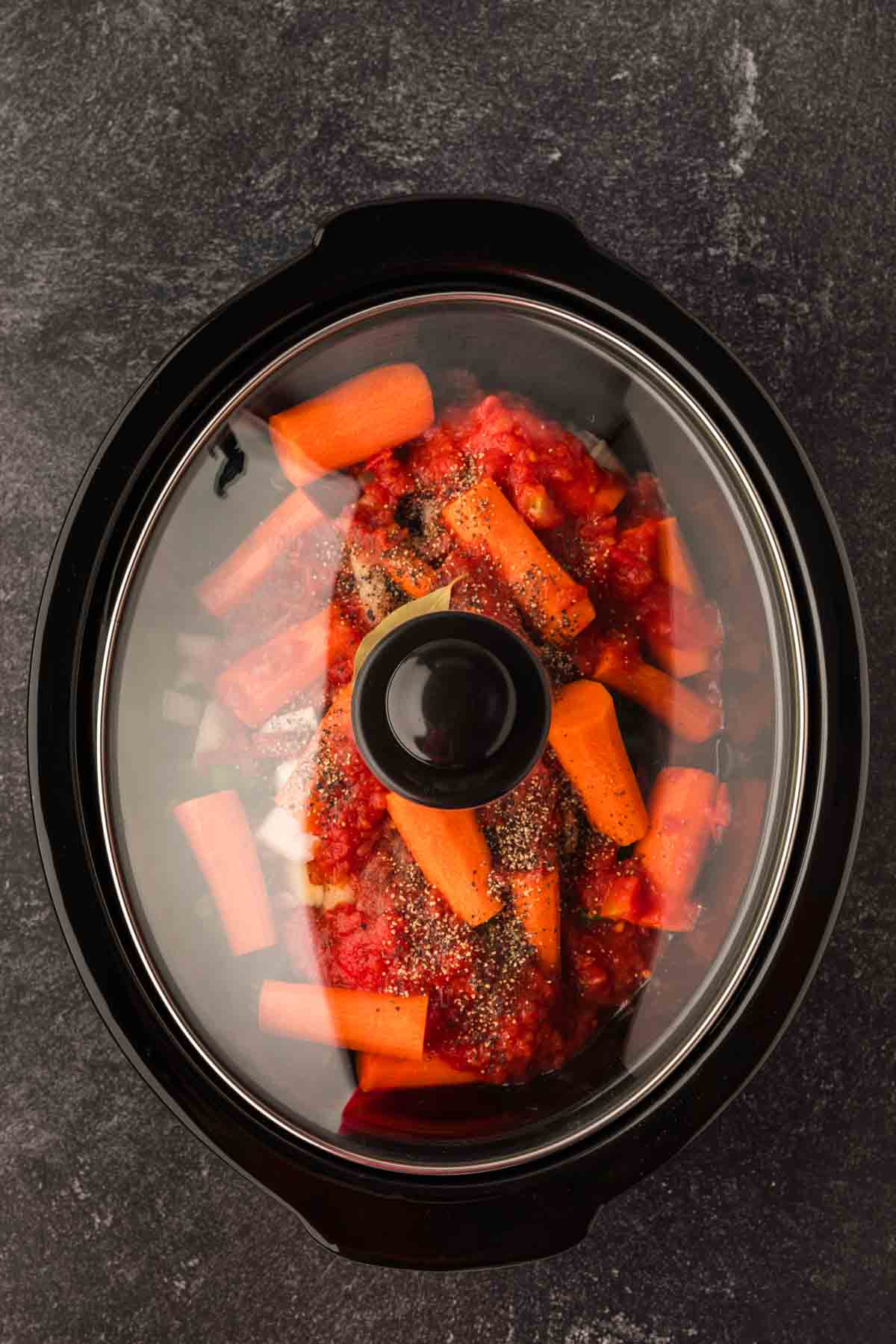 A slow cooker filled with chopped carrots, onions, tomatoes, and seasonings, just right for a Crock Pot beef stew recipe. It has a black exterior and a glass lid that reveals the colorful ingredients inside. The cooker sits on a dark countertop, ready to transform these components into culinary delight.