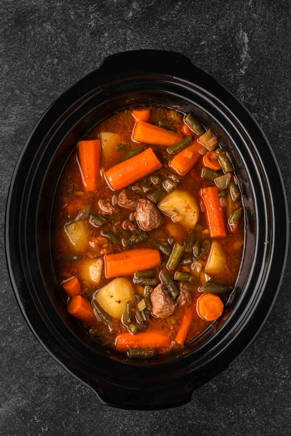 A black slow cooker showcases a delicious Crock Pot Beef Stew Recipe, with tender chunks of beef, carrots, potatoes, and green beans all mingling in a rich broth. The ingredients are beautifully mixed and visible from above.