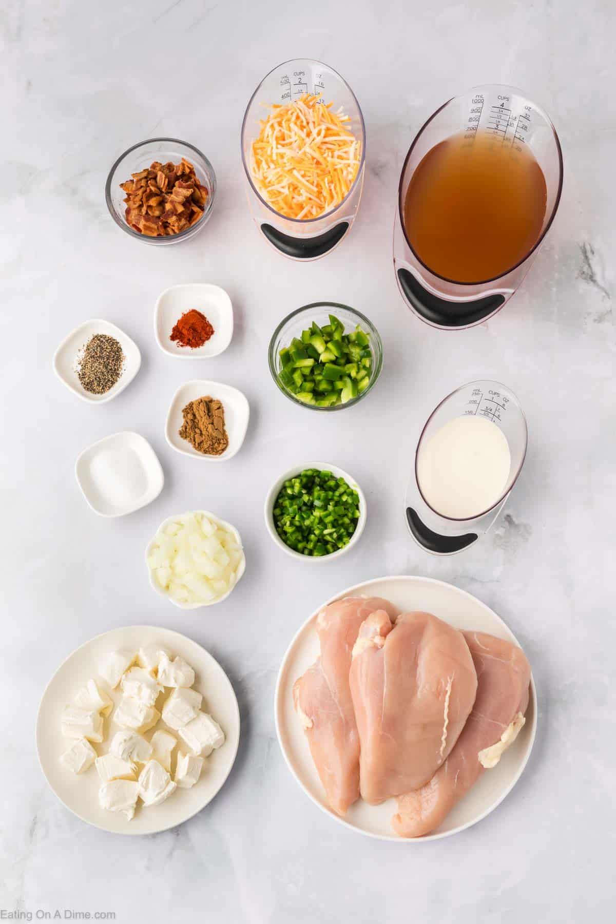 Ingredients for Crock Pot Jalapeño Popper Chicken Soup Recipe on a marble surface: raw chicken breasts, cream cheese cubes, chopped onion, diced green peppers, minced green chili, shredded cheese, chicken broth, heavy cream, bacon bits, and various spices in bowls and measuring cups.