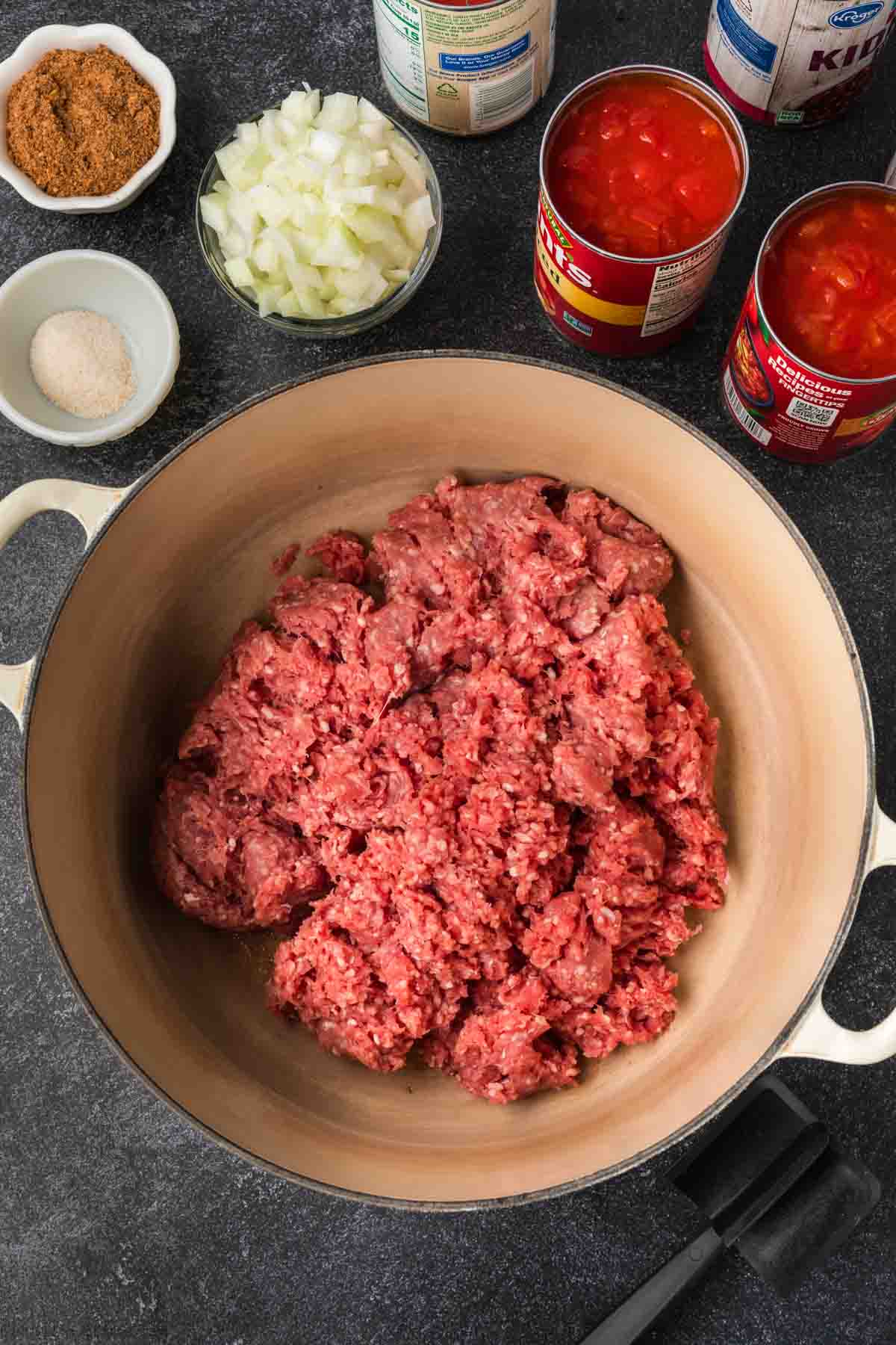 A pot with ground meat surrounded by canned tomatoes, chopped onions, and a small bowl of seasoning and spices sits invitingly on a dark countertop—a perfect setup for an easy chili recipe.