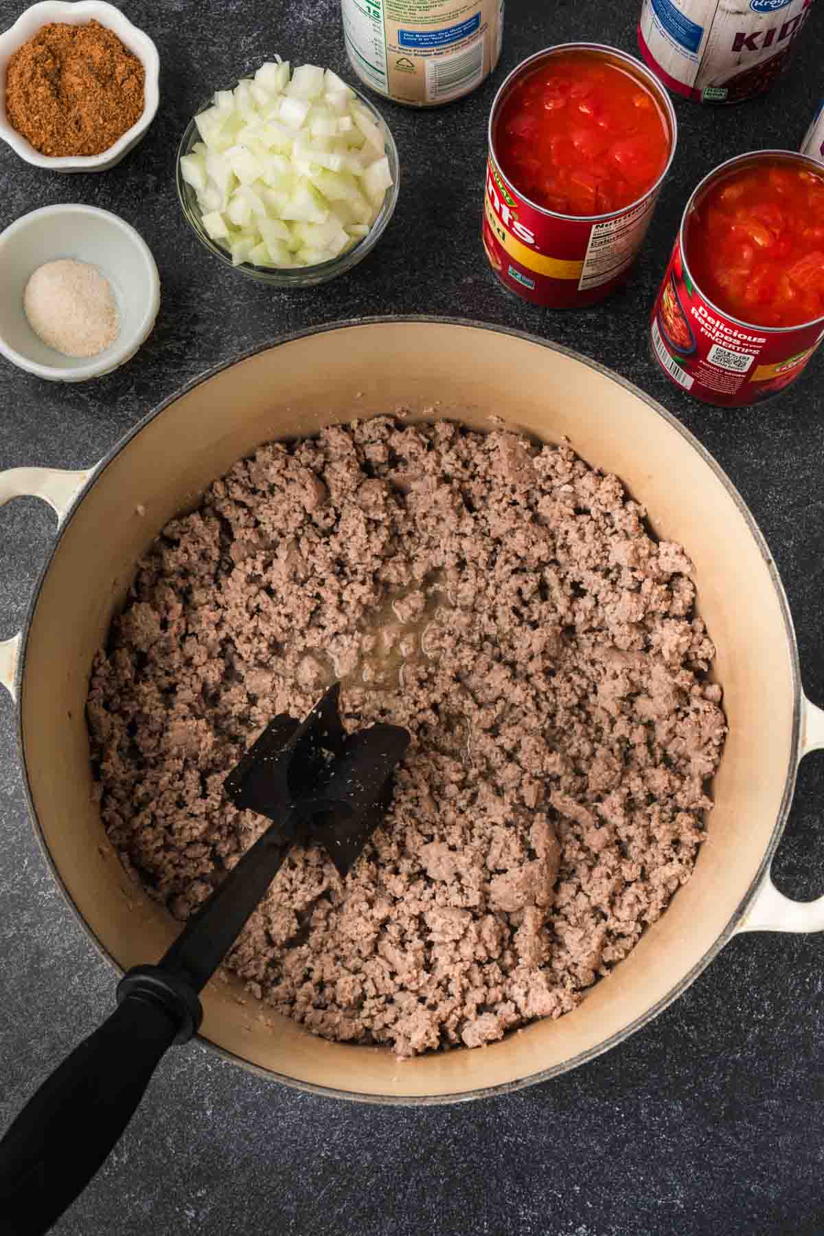 A pot of browned ground beef sits on a stovetop with a black spatula, hinting at an easy chili recipe in the making. Surrounding the pot are diced onions in a bowl, canned tomatoes, and various small containers of spices, all ready to come together for a hearty dish.