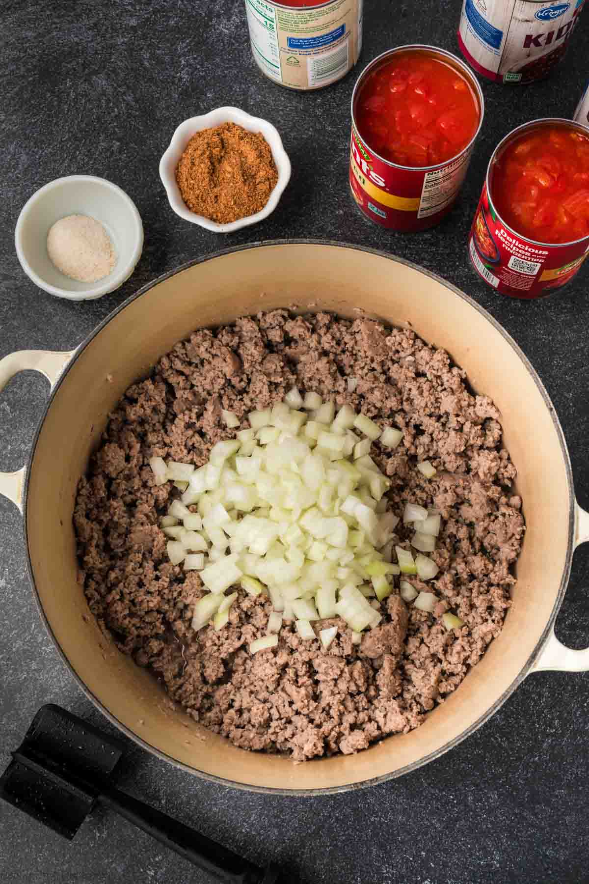 A pot filled with browned ground meat topped with chopped onions invites you to try this easy chili recipe. Surrounding the pot are cans of peeled tomatoes, kidney beans, and bowls containing spices. A meat masher is placed nearby on a dark surface, ready for action.