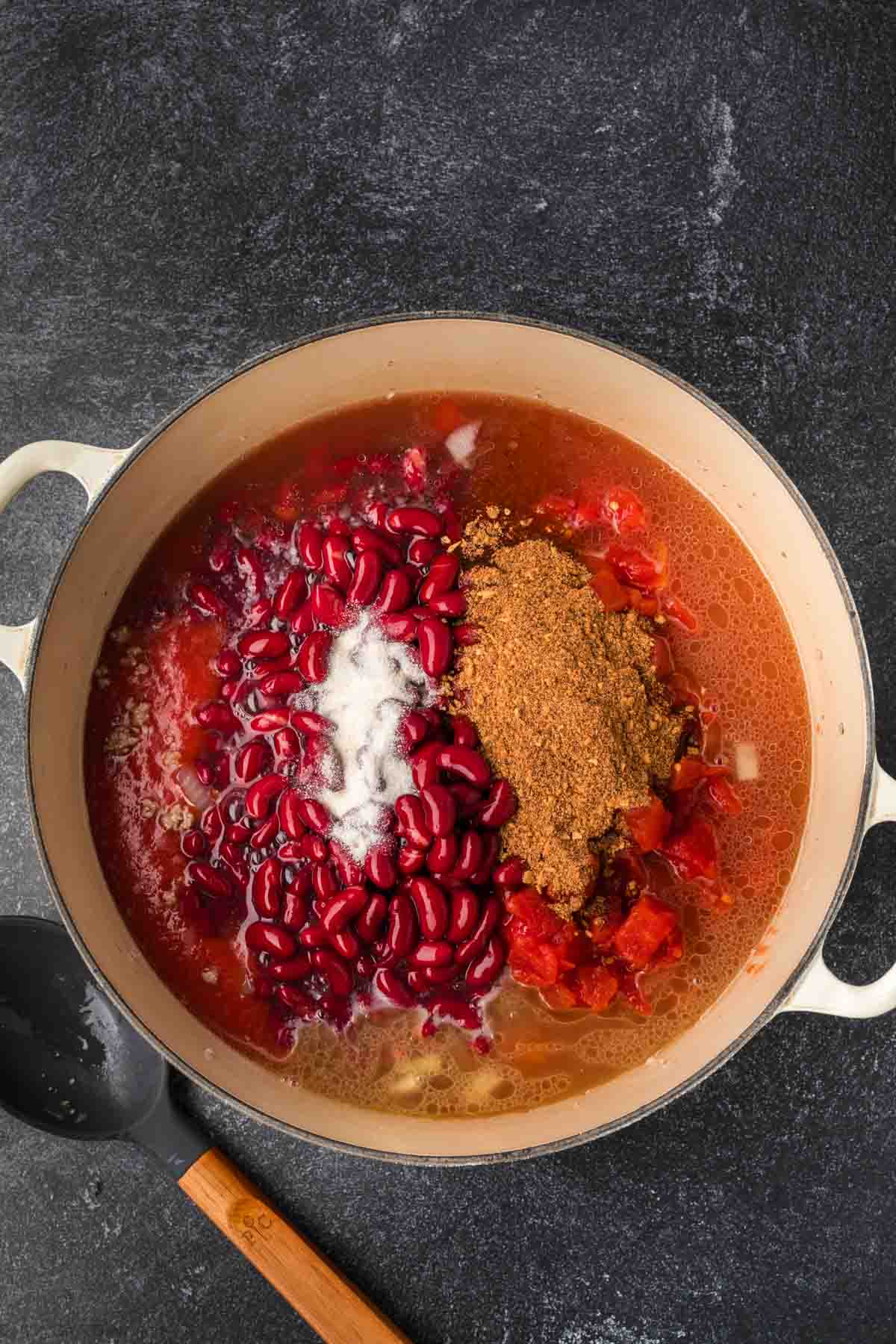 A pot brimming with ingredients for an easy chili recipe, featuring red kidney beans, chopped tomatoes, spices, and a seasoning mix on a dark countertop. A black spoon with a wooden handle rests beside the pot.