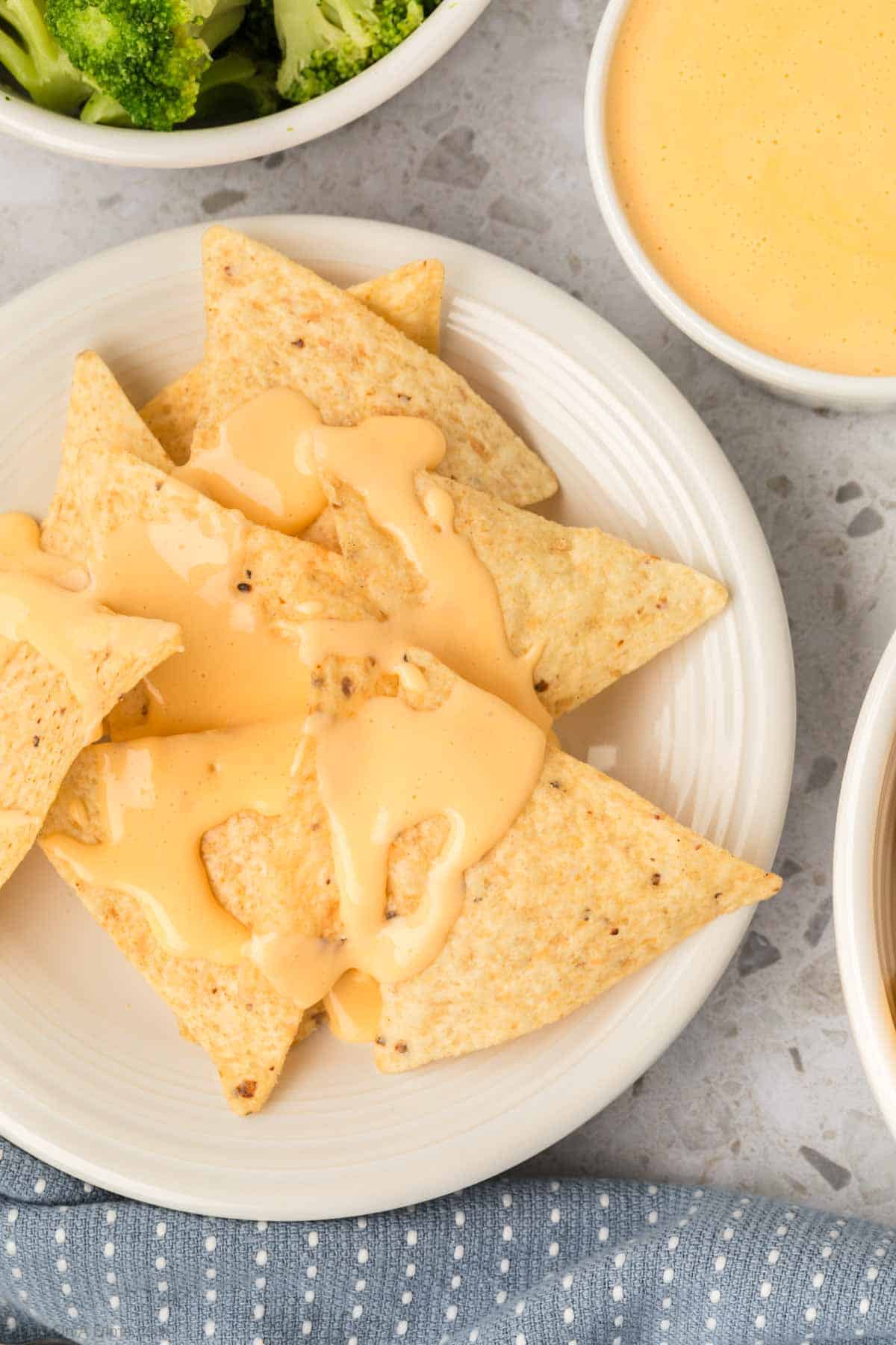 A plate of tortilla chips drizzled with creamy American cheese sauce sits on a light grey countertop. Nearby, a small bowl of cheese sauce and a dish with broccoli florets accompany the snack. A textured blue napkin is partially visible in the foreground.