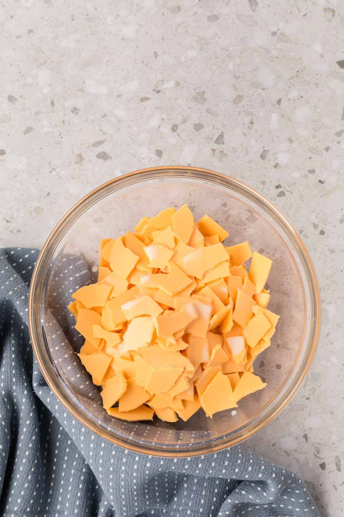 A glass bowl filled with crumbled yellow cheese, perfect for creating a smooth American Cheese Sauce, sits on a gray speckled countertop. A blue cloth with white dots is partially tucked under the bowl, adding a touch of charm to the scene.