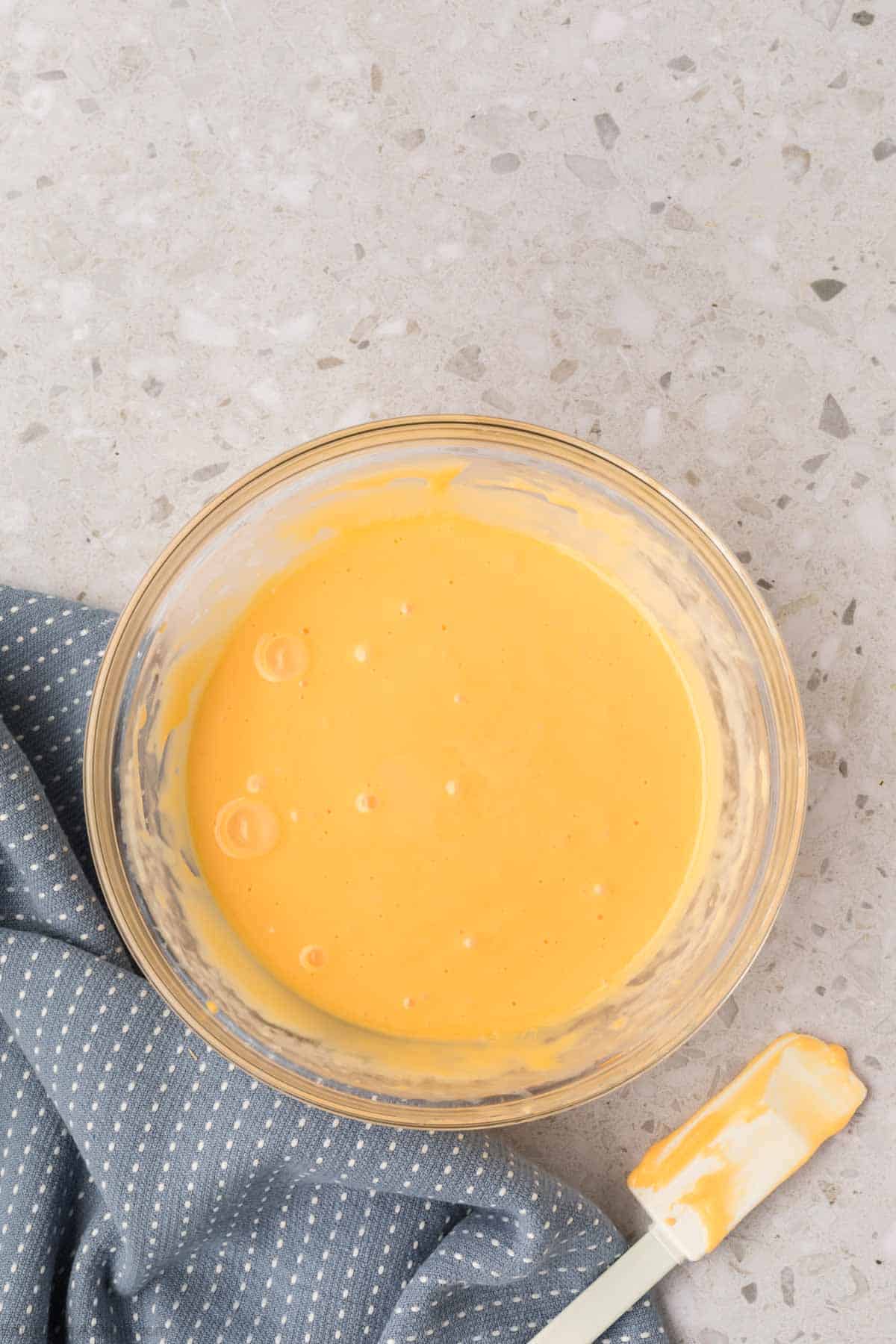 A bowl of smooth, creamy orange batter reminiscent of American cheese sauce sits on a speckled countertop, next to a white spatula with batter remnants. A blue cloth with small white dots is folded beside the bowl.