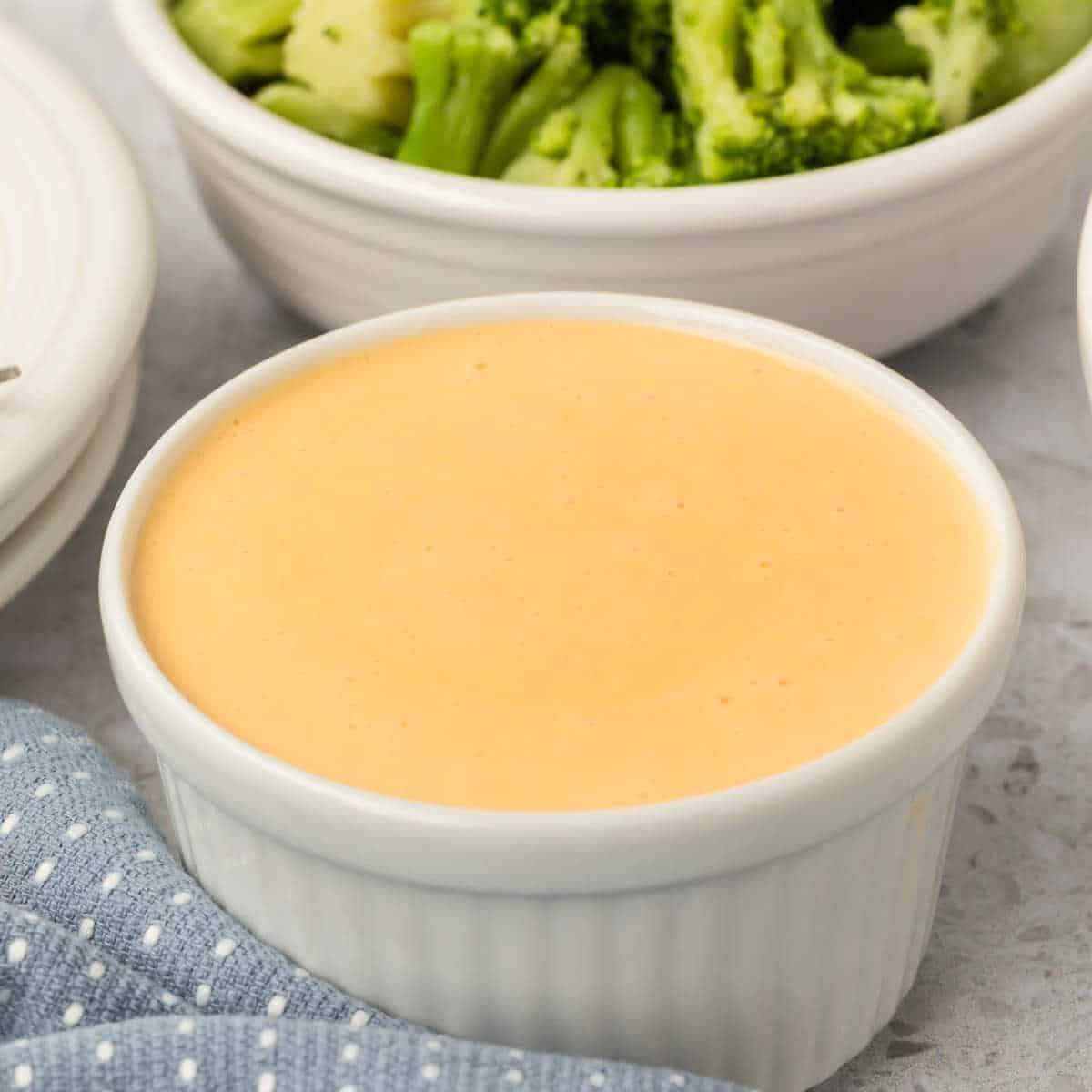 A small white bowl filled with creamy American cheese sauce sits on a white surface. In the background, a bowl of steamed broccoli and part of a blue cloth with white polka dots add a burst of color.