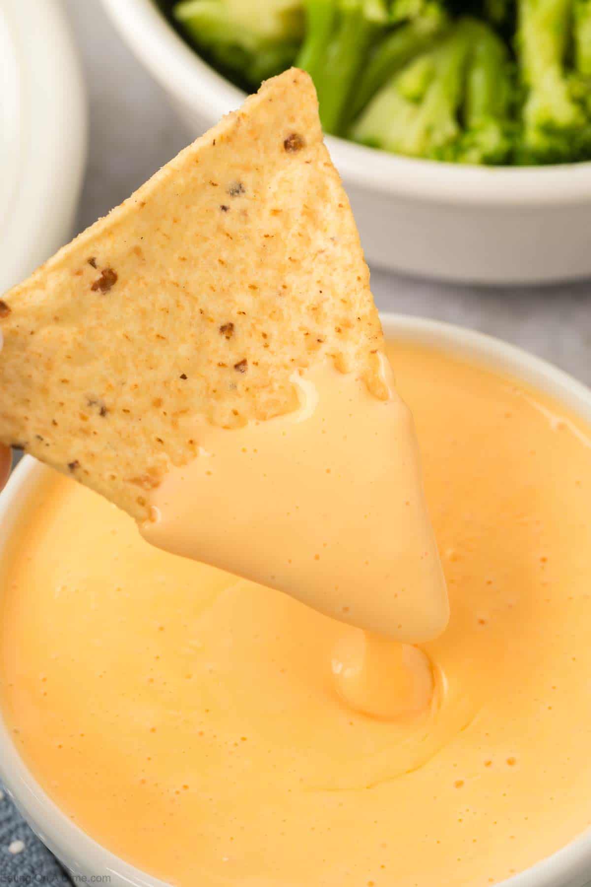 A nacho chip dives into a creamy orange pool of American cheese sauce. In the background, a bowl of green broccoli adds a splash of color.