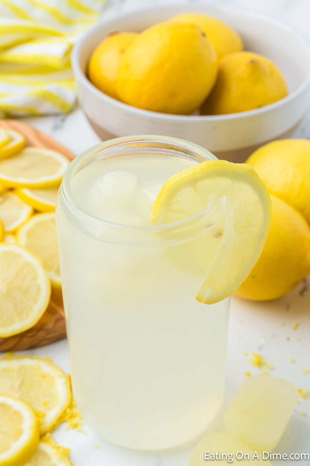 Glass of lemonade with a bowl of whole lemons and lemon slices on the side