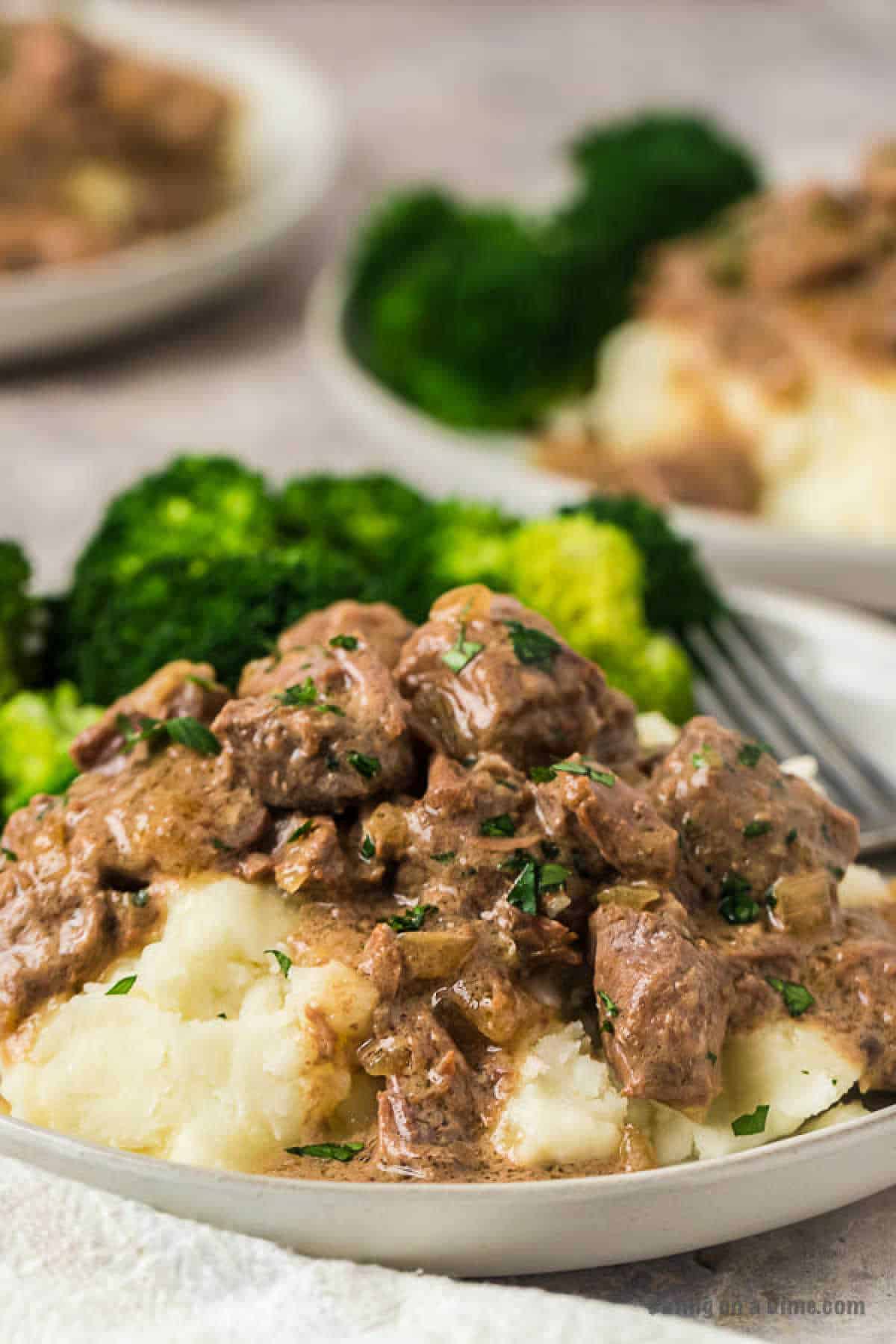 A plate of mashed potatoes topped with creamy Instant Pot beef tips and gravy, garnished with herbs. In the background, there are florets of steamed broccoli. The dish is served on a white plate.