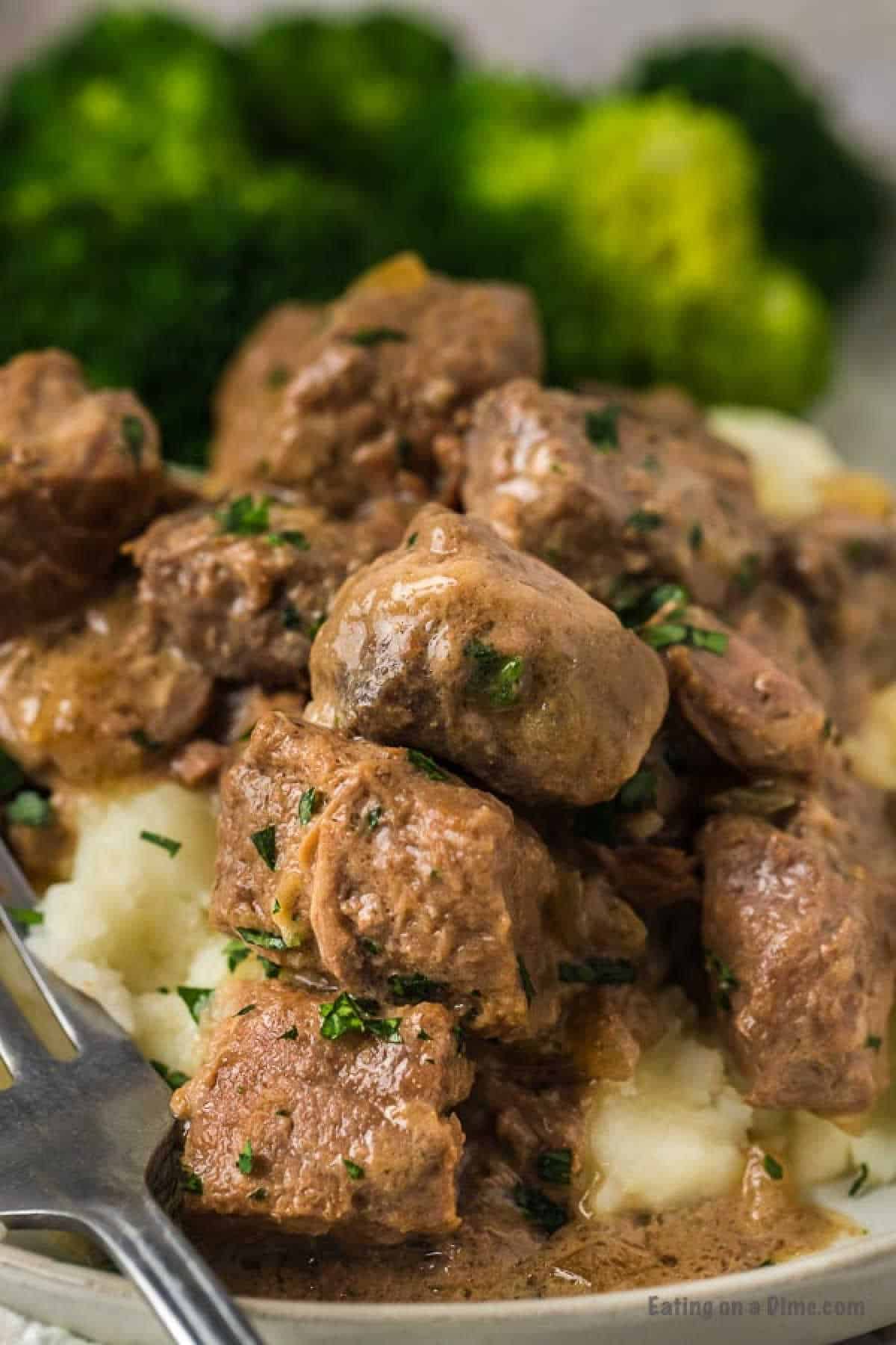 A plate of tender Instant Pot beef tips and gravy garnished with fresh herbs, served over a bed of creamy mashed potatoes with a side of steamed broccoli. A fork is placed on the plate, ready for eating.