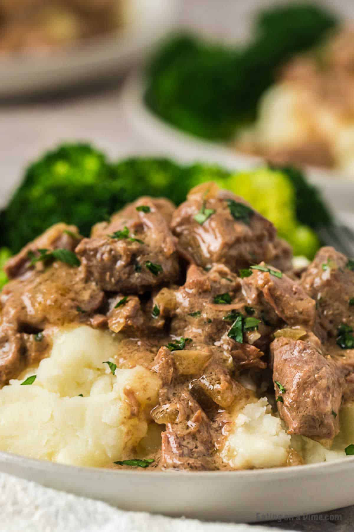 A plate of creamy mashed potatoes topped with Instant Pot beef tips and gravy, garnished with chopped herbs. In the background, there is a serving of steamed broccoli, adding a pop of green to the dish.