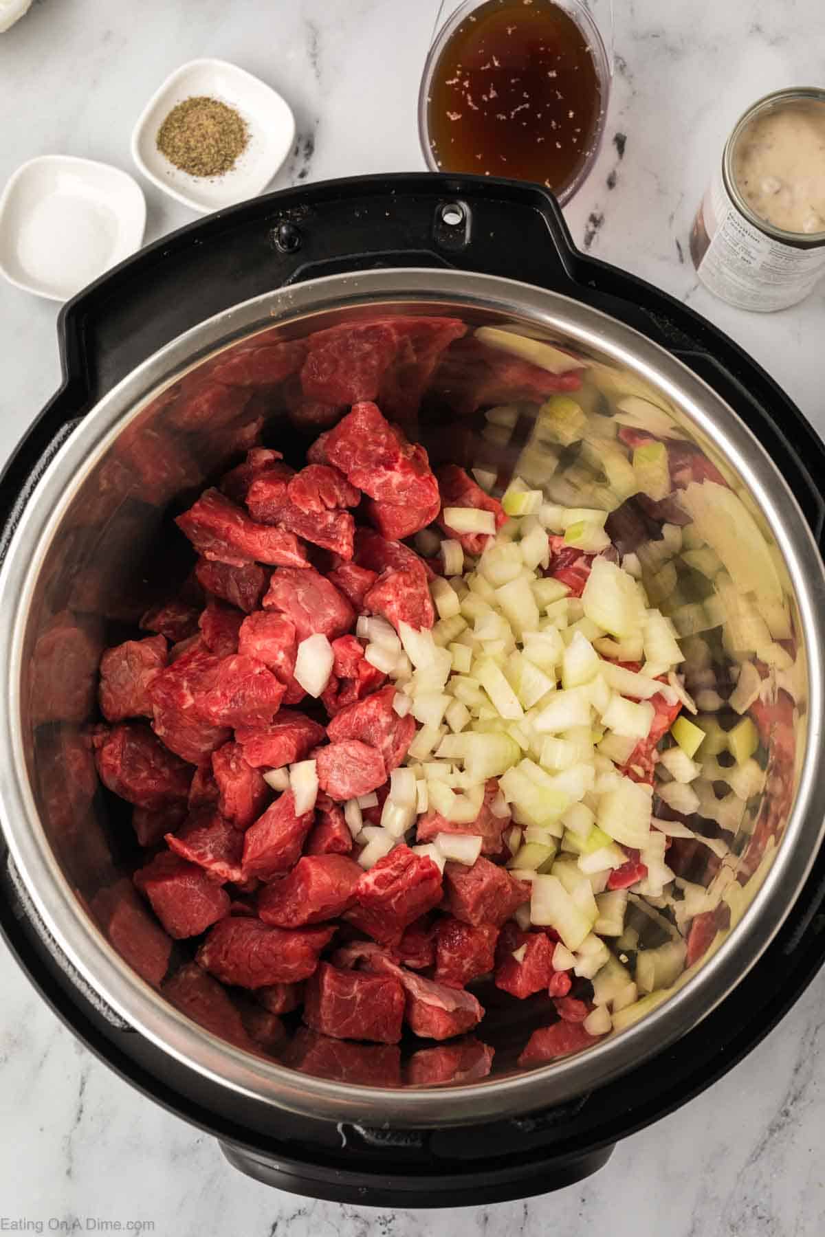 An open pressure cooker filled with raw beef chunks and chopped onions hints at a savory meal in the making. Instant Pot Beef Tips and Gravy ingredients such as pepper and broth await nearby in small bowls on the marble countertop.